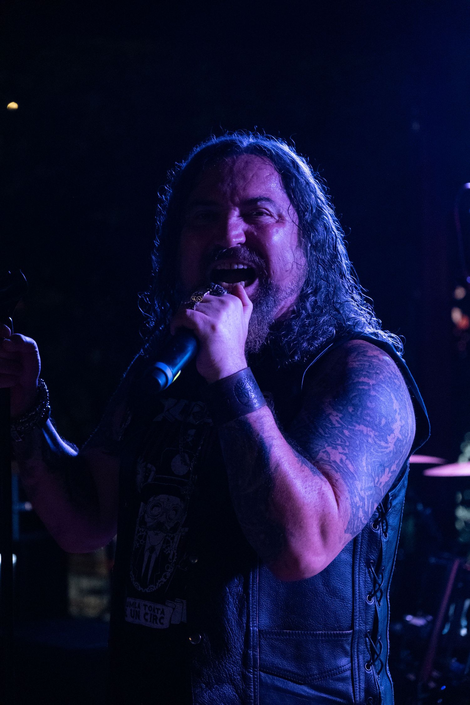 Long-haired male rock vocalist with tattoos singing into microphone under purple stage lights.