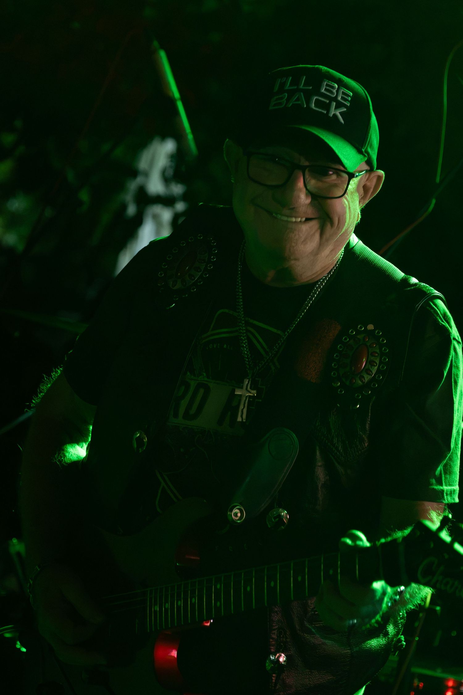 Smiling guitarist wearing a cap and cross necklace performing on stage under green lights.