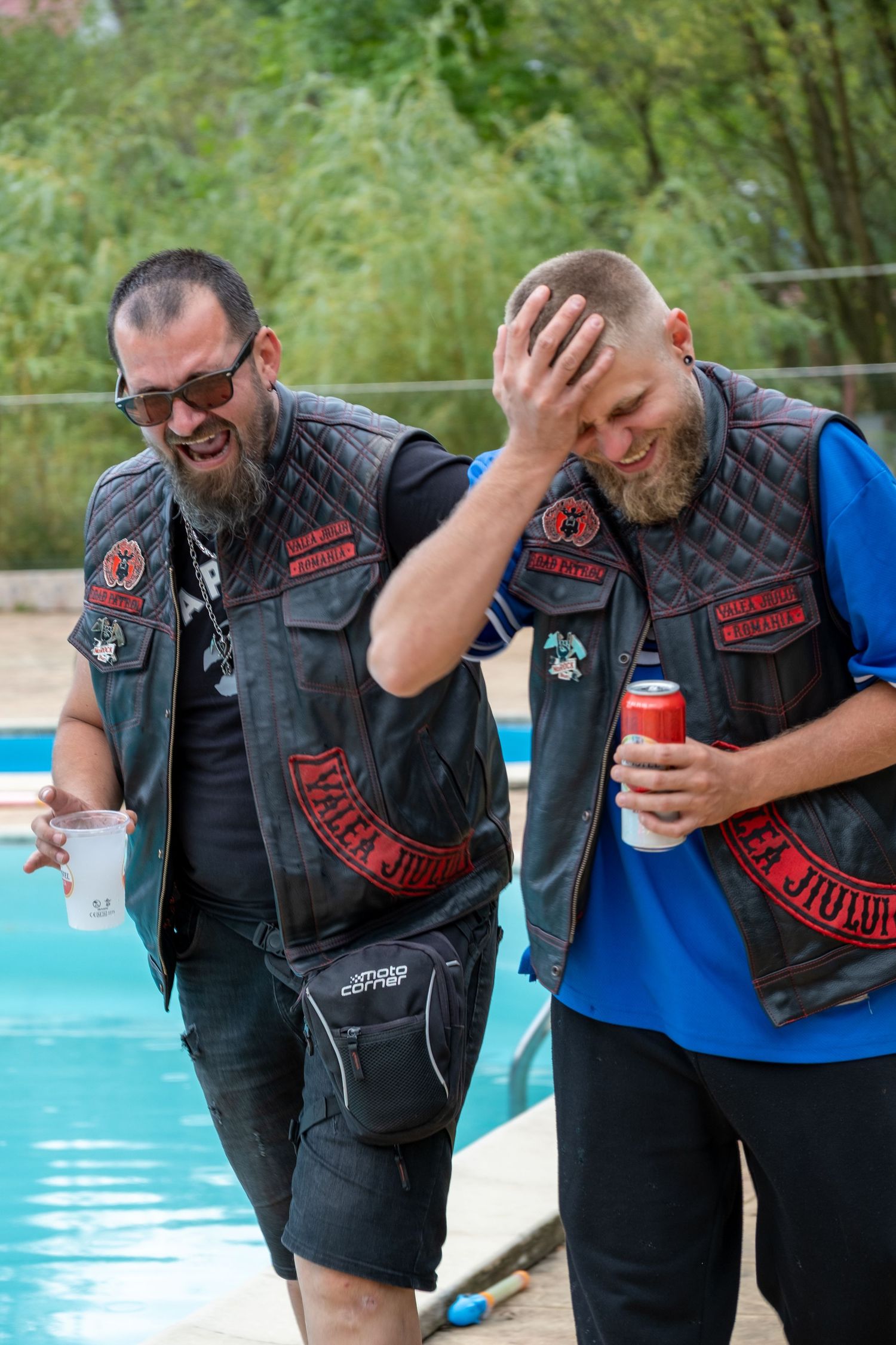 Two laughing men in biker leather vests with patches standing near a swimming pool.