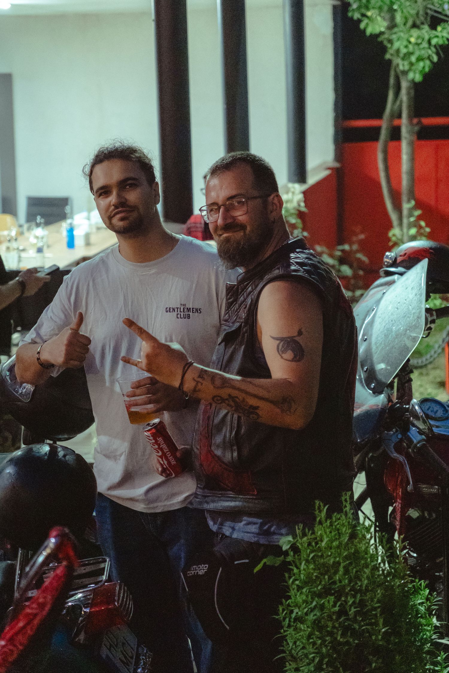 Two men pose with motorcycles at a biker club event, one flashing a rock sign while holding a beer.