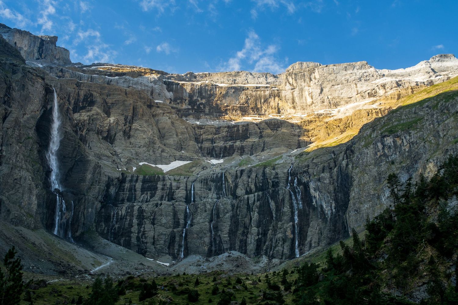 Dramatic mountain cirque with a tall waterfall cascading down rocky cliffs under blue sky.