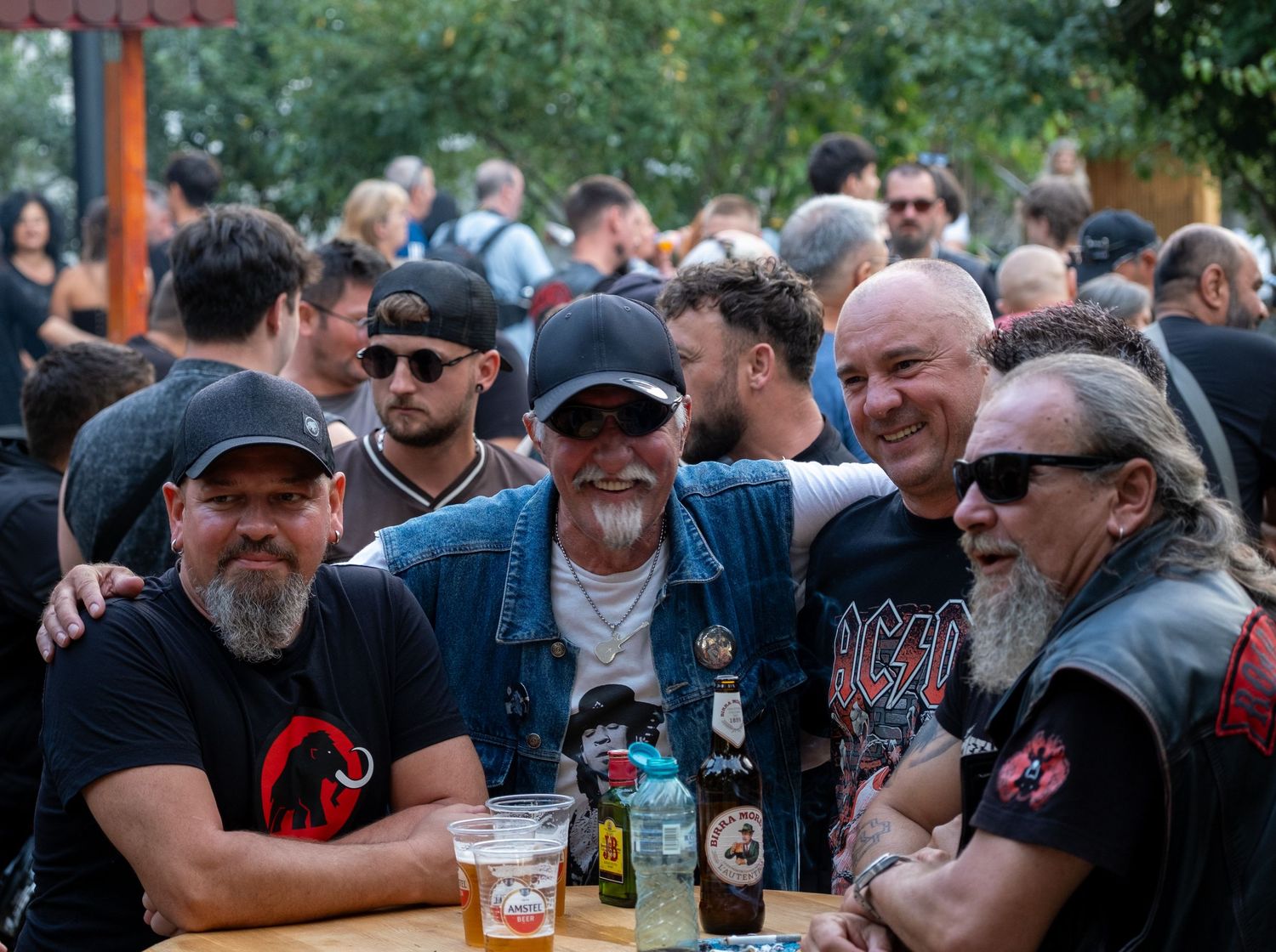 Group of five smiling biker men posing together at an outdoor motorcycle rally event.