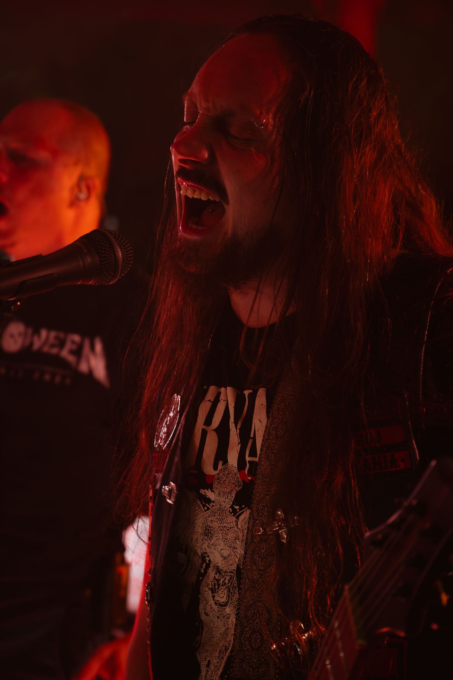 Long-haired metal vocalist screams into microphone under dramatic red stage lighting at concert.