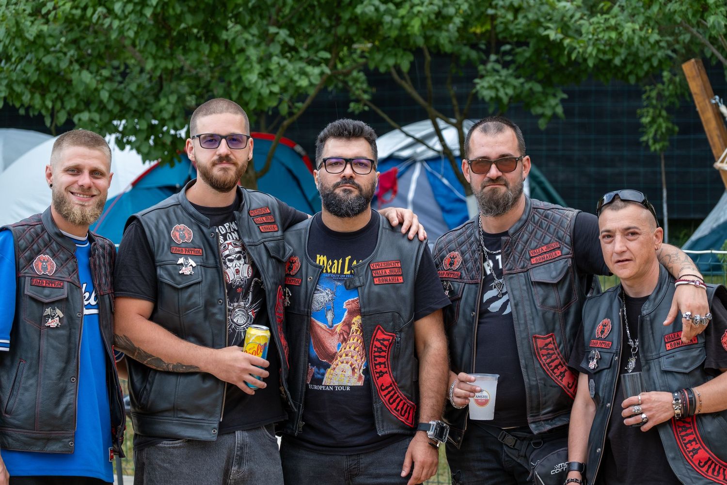 Five men in biker vests with patches pose together outdoors at a motorcycle event.