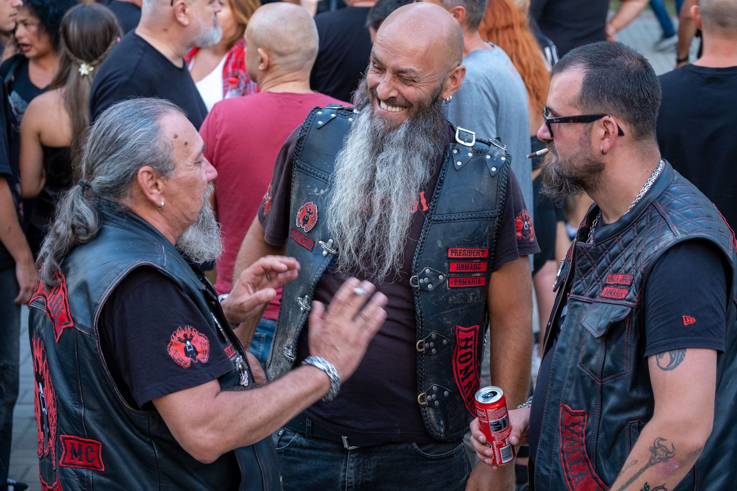 Three biker men in leather vests laughing and conversing at an outdoor motorcycle club event.