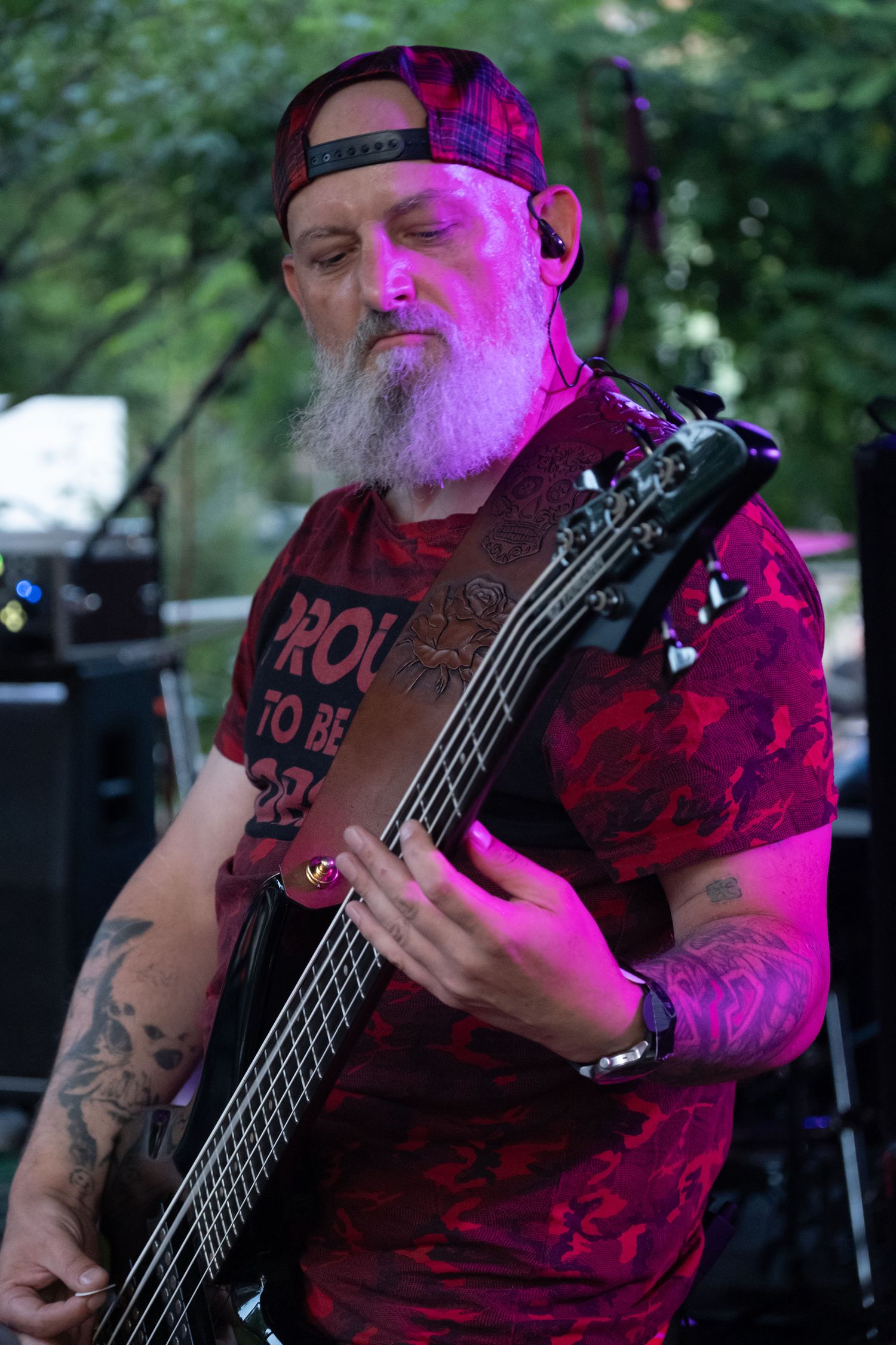 Tattooed male bassist with white beard and red cap performs on stage under purple lights.