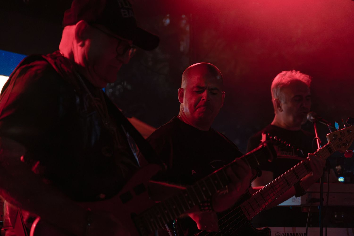 Three guitarists perform together under red stage lighting at an intimate rock concert.