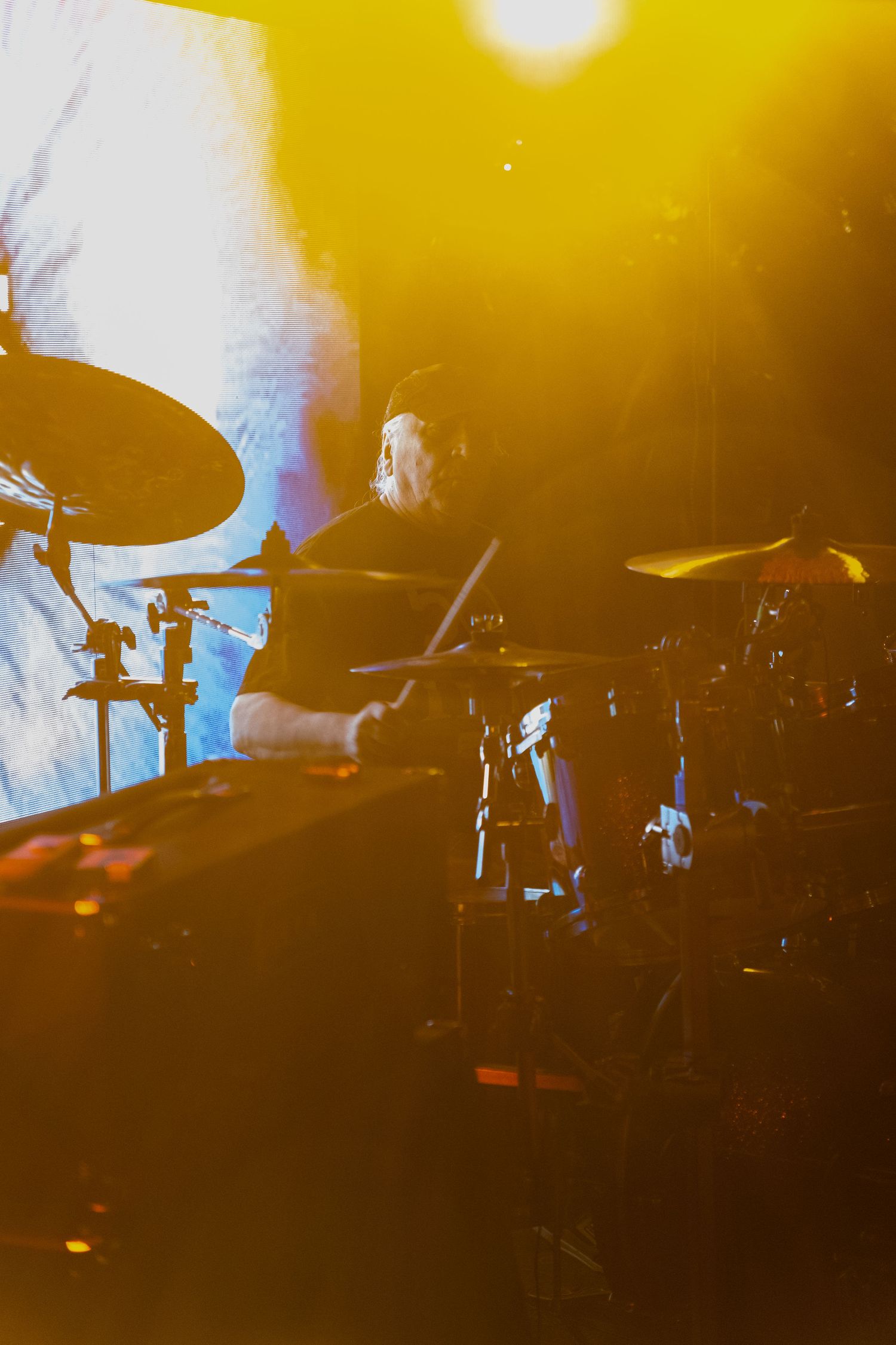 Drummer performing on stage under warm golden spotlight with large projection screen behind.