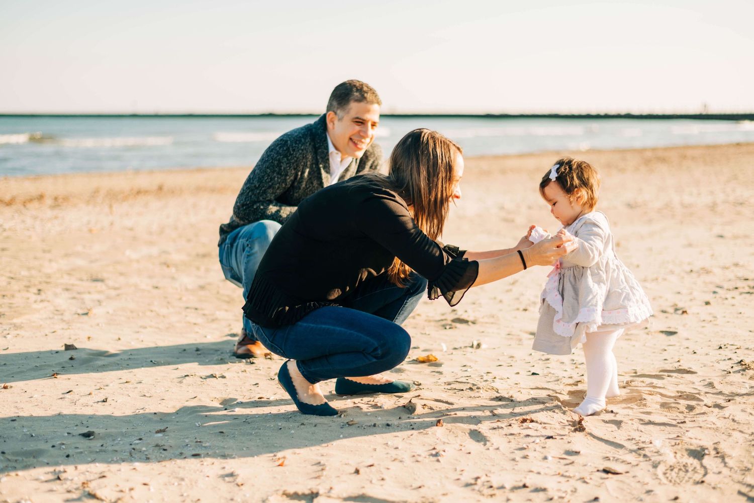 mother helping her baby girl walk while father looks on at the beach