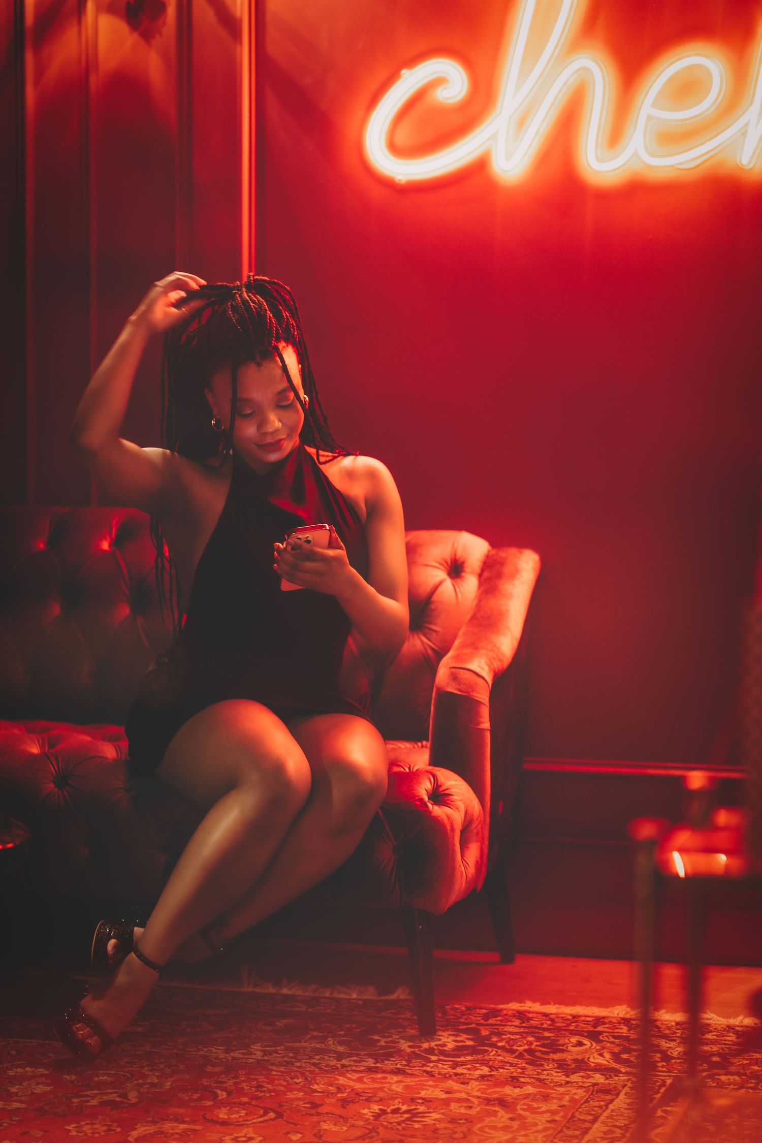 Woman in black dress sitting in red-lit lounge chair with neon sign.