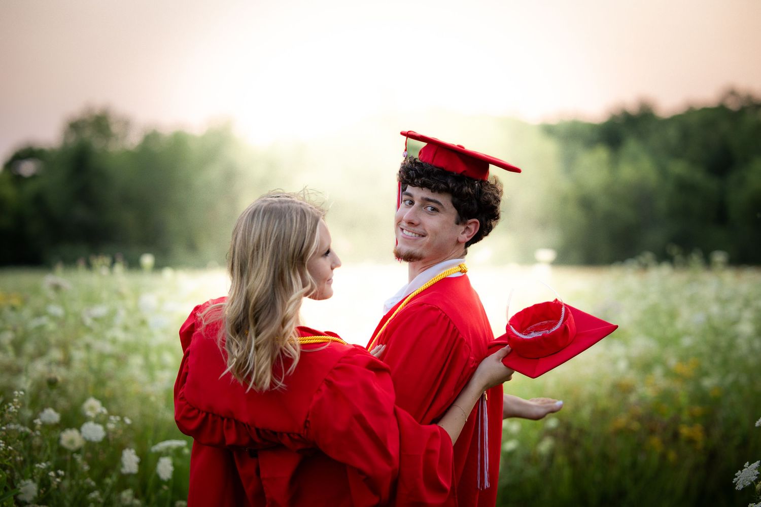 Two graduates in red caps and gowns share a romantic moment in a sunlit wildflower field.