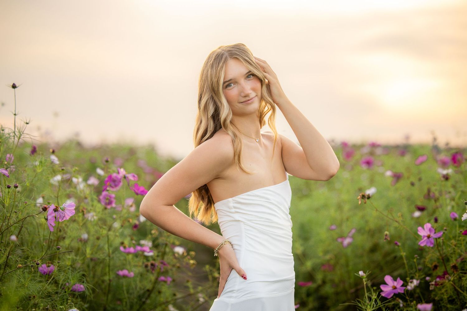 Young blonde woman in white dress posing in a vibrant pink flower field at golden hour sunset.