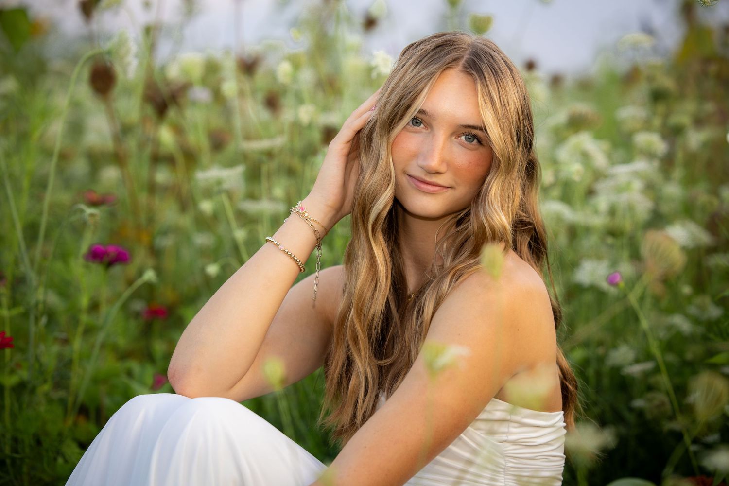 Young woman with long blonde hair sitting in a wildflower field wearing a white dress, smiling softly.