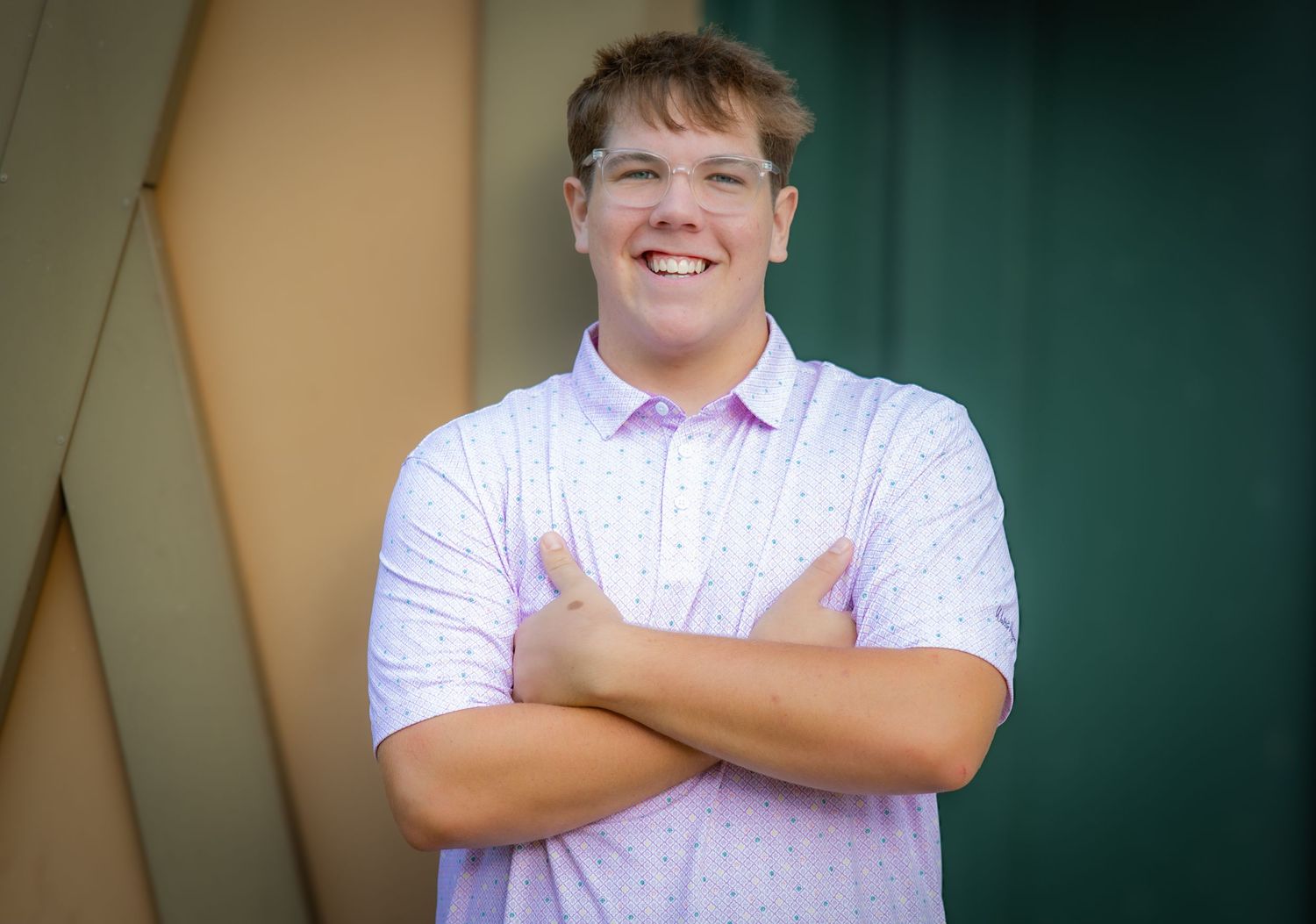 Smiling young man with arms crossed wearing a light purple polo shirt, posing outdoors near a green wall.