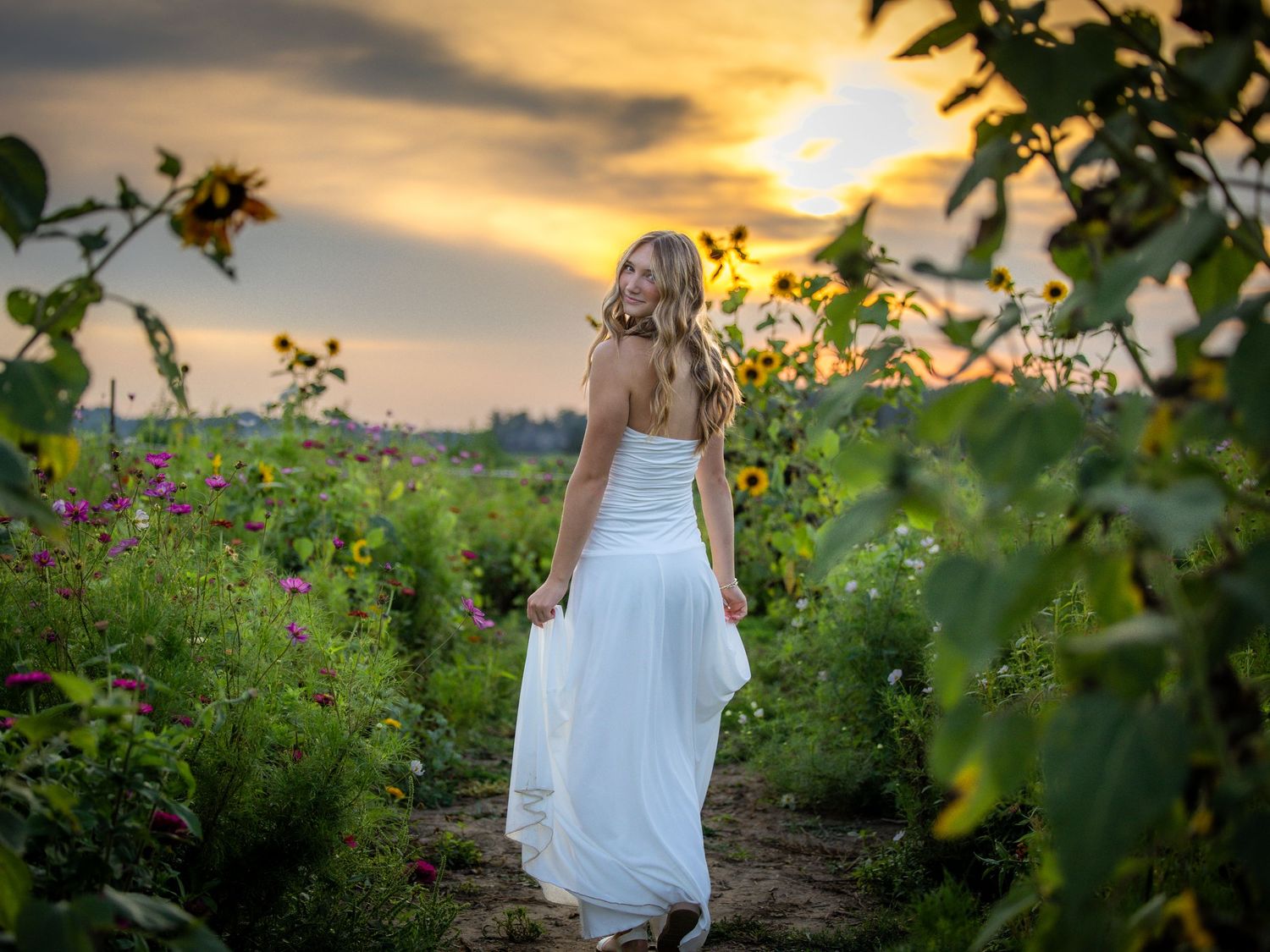 Woman in white dress walking through a vibrant flower garden at golden sunset.