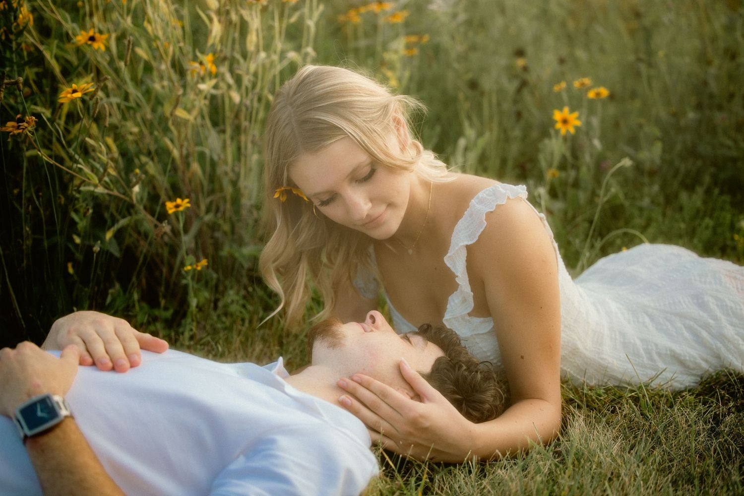 A blonde woman in a white dress tenderly gazes at a young man lying in a flower field.