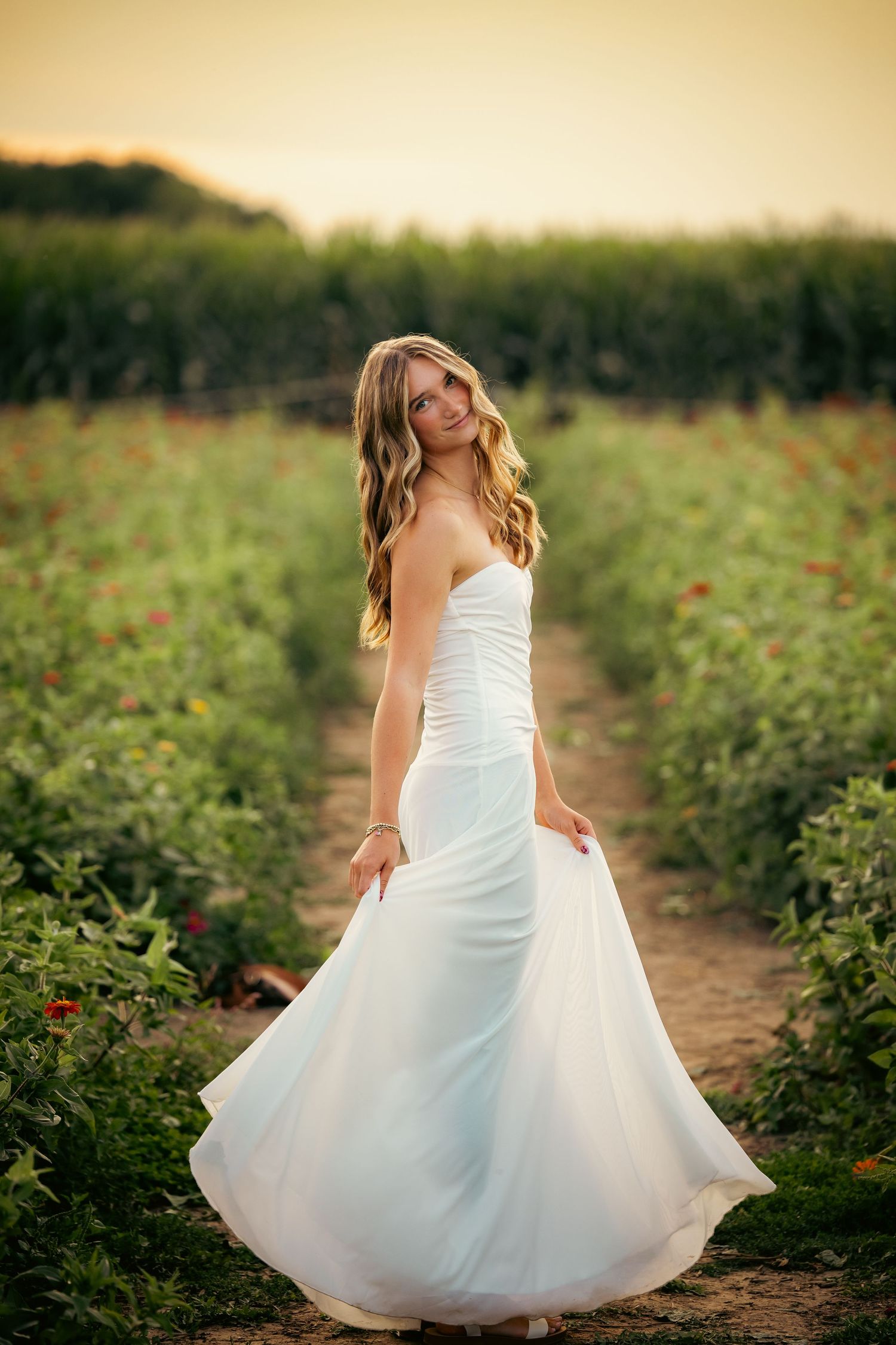 A bride in a flowing white strapless gown twirls in a lush green field at golden hour.