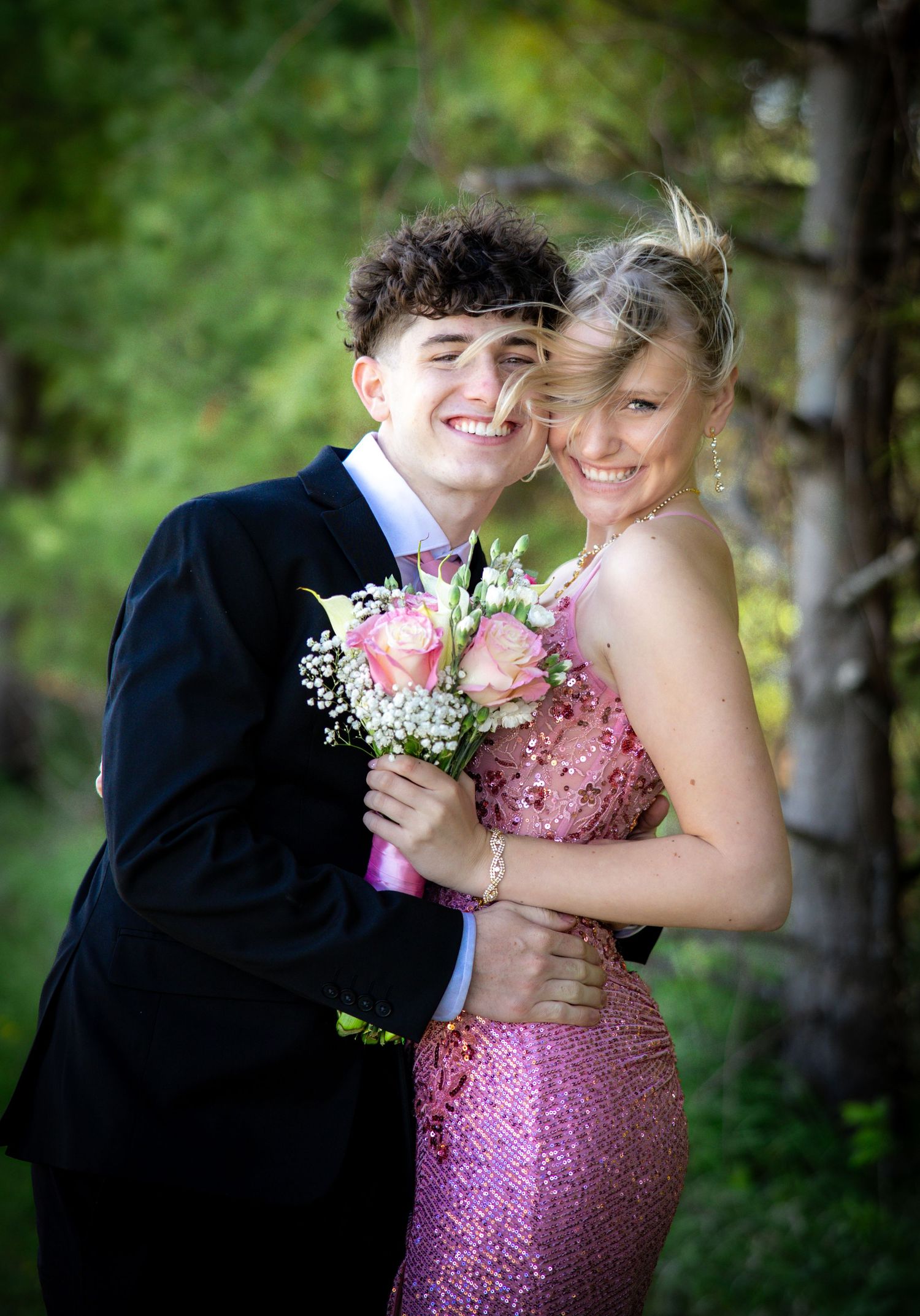 Smiling teenage prom couple posing outdoors with pink rose bouquet in forest setting.