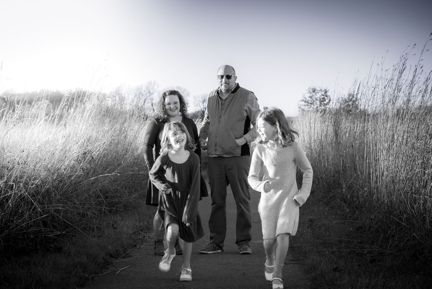 A joyful family of four walks together along a scenic outdoor path in a black and white photo.