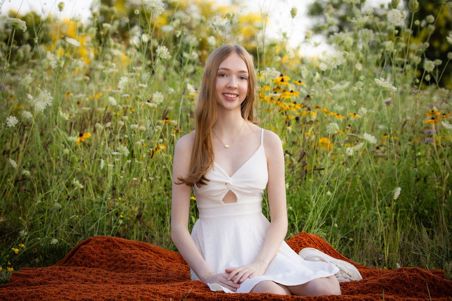 Young woman in white sundress sitting on orange blanket surrounded by wildflowers in a lush green meadow.