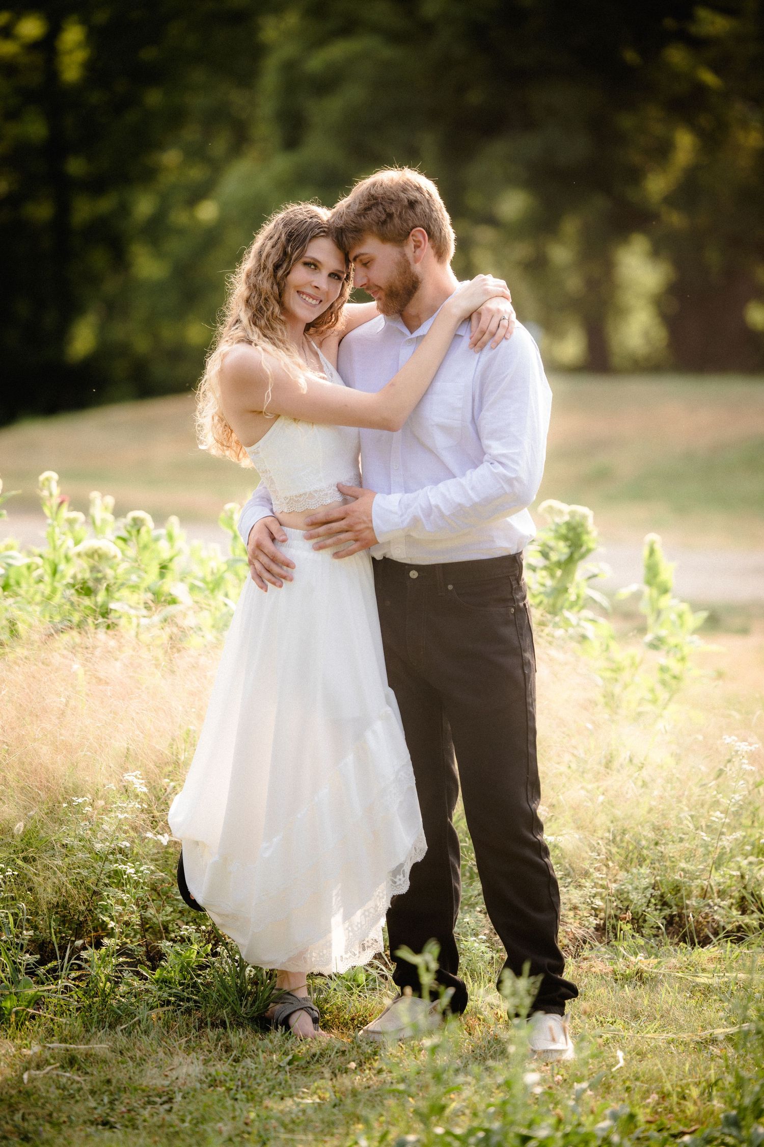 Couple embracing in sunlit park, bride in white dress and groom in white shirt, sharing a romantic outdoor moment.