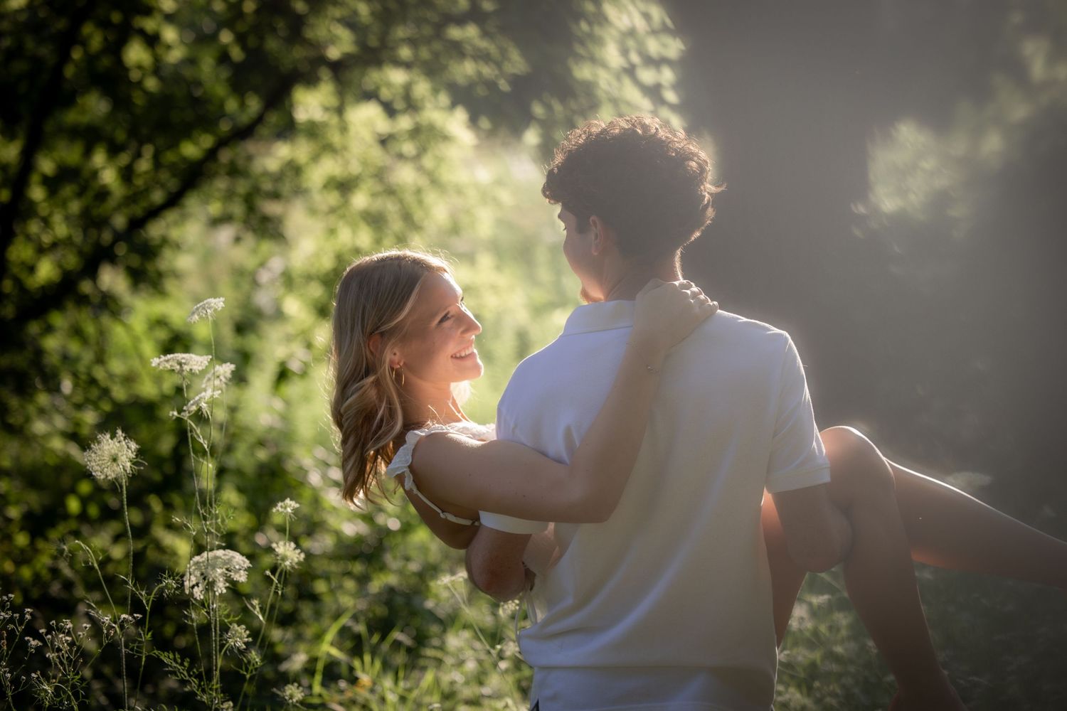 A couple embraces and smiles at each other in a sunlit garden surrounded by lush green foliage and white wildflowers.