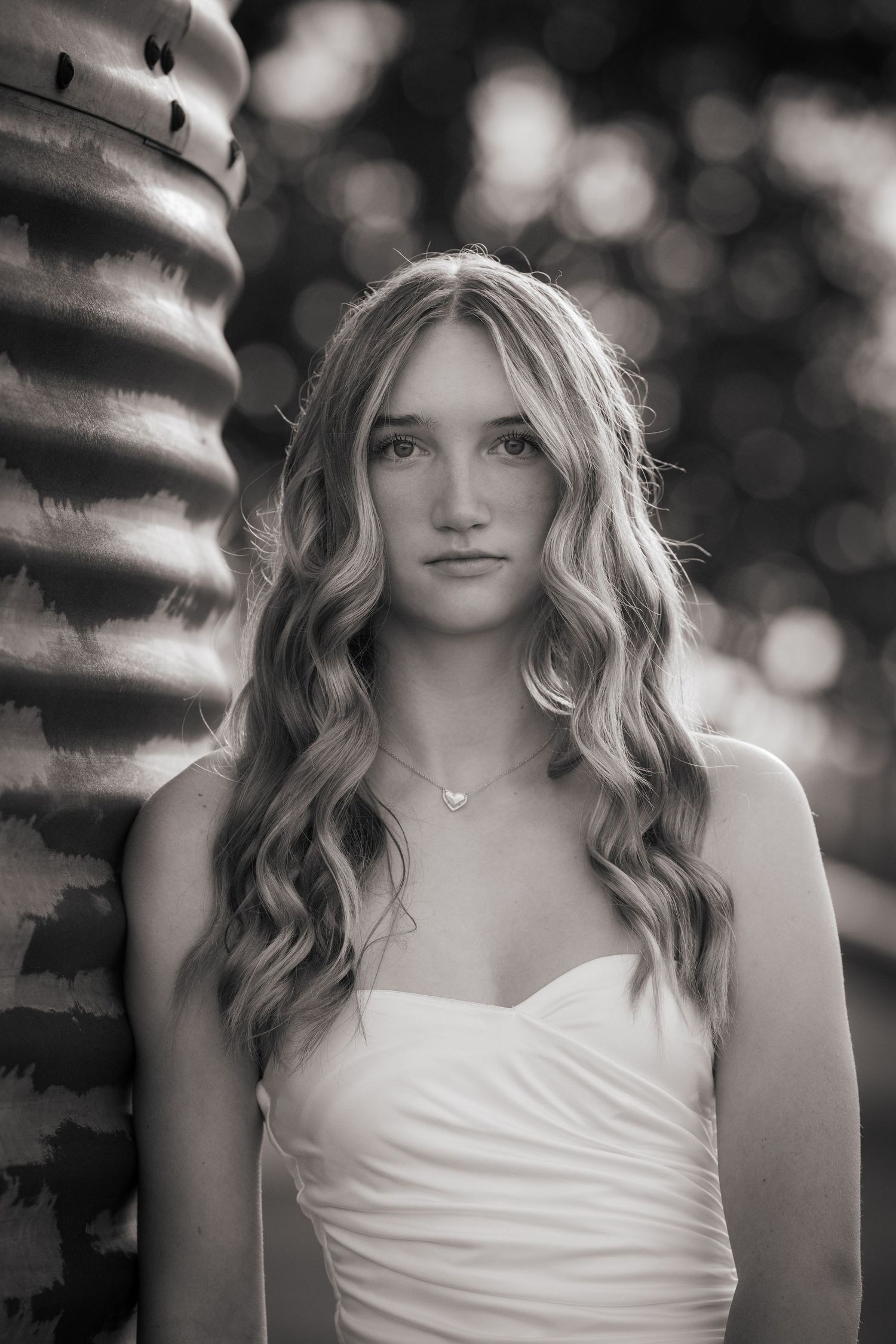 Black and white portrait of a young woman with long wavy blonde hair wearing a white strapless dress and delicate necklace.