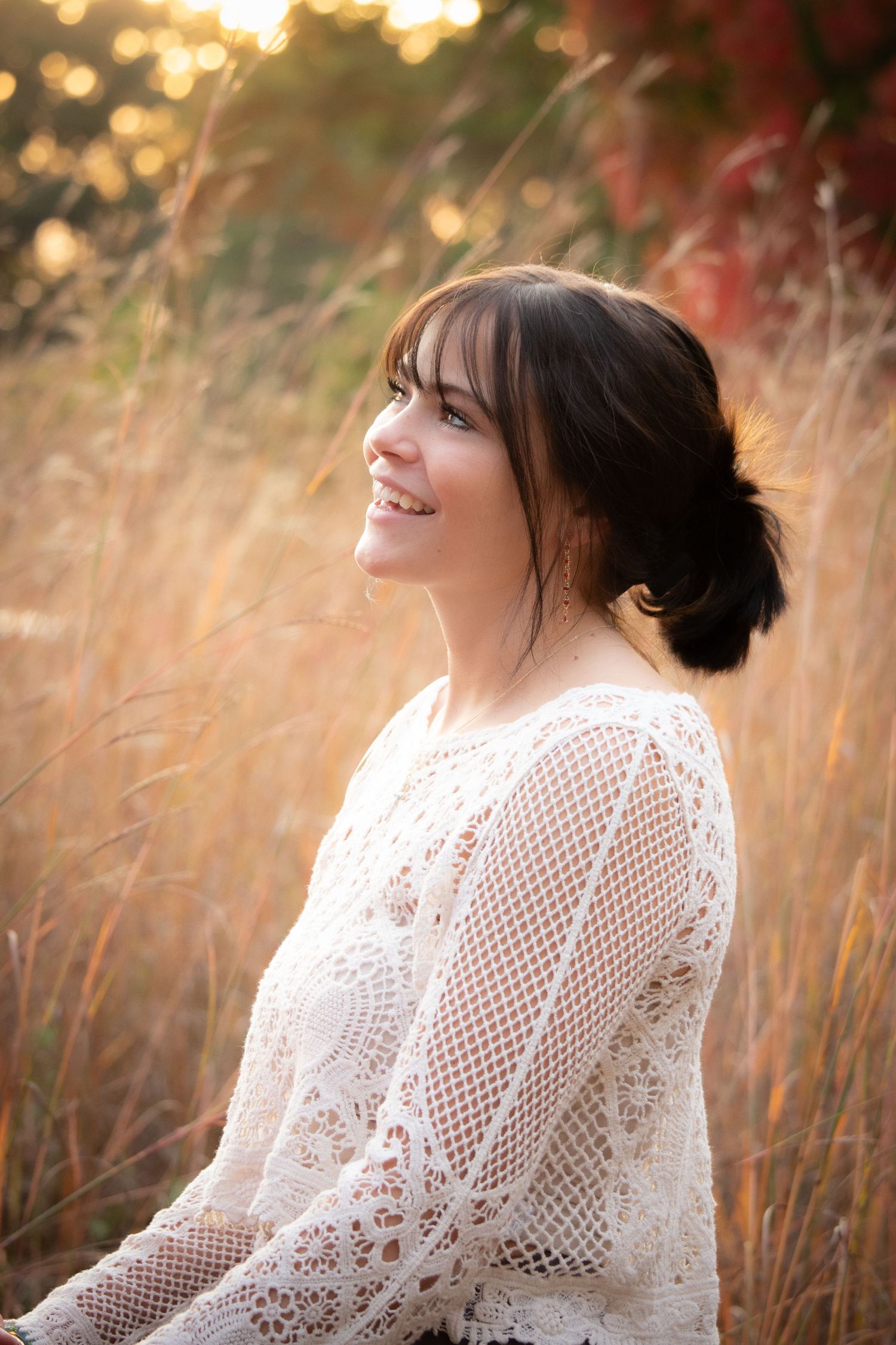 Smiling brunette woman in white lace top sitting in golden grass field during sunset portrait.