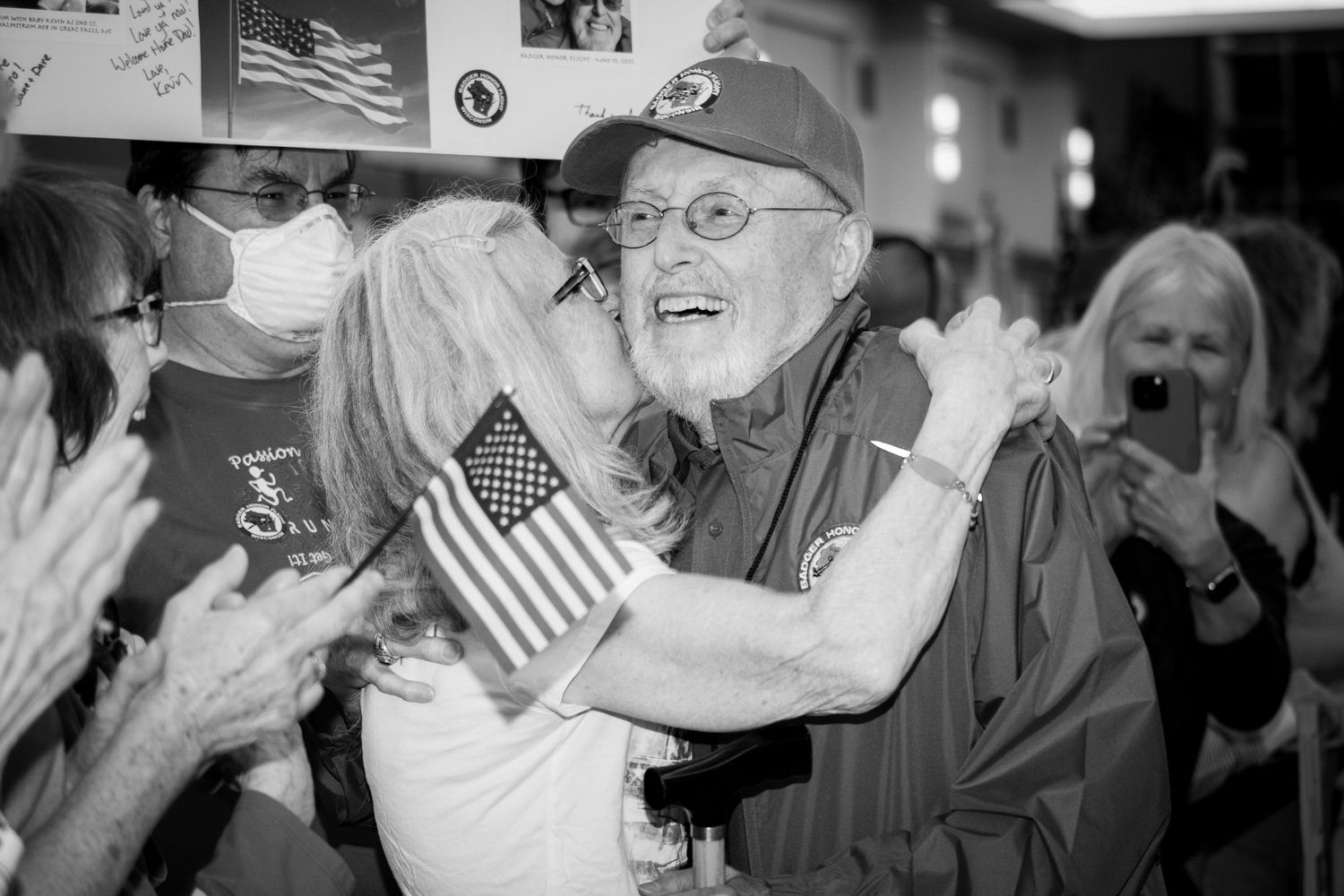 Elderly man receiving a kiss on the cheek at a patriotic celebration holding an American flag.
