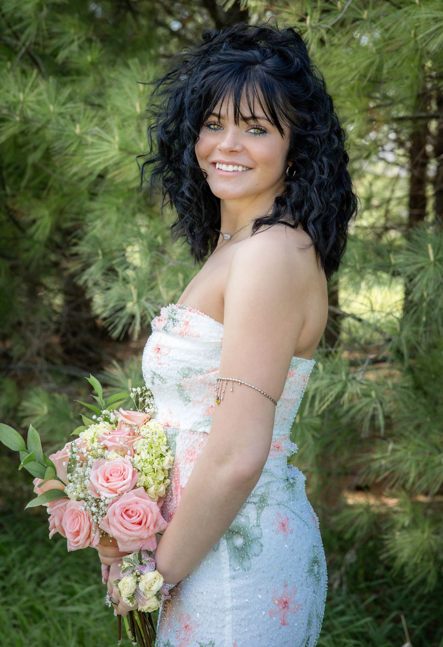 Smiling Senior with dark curly hair holding pink roses bouquet, wearing strapless floral gown outdoors among pine trees.