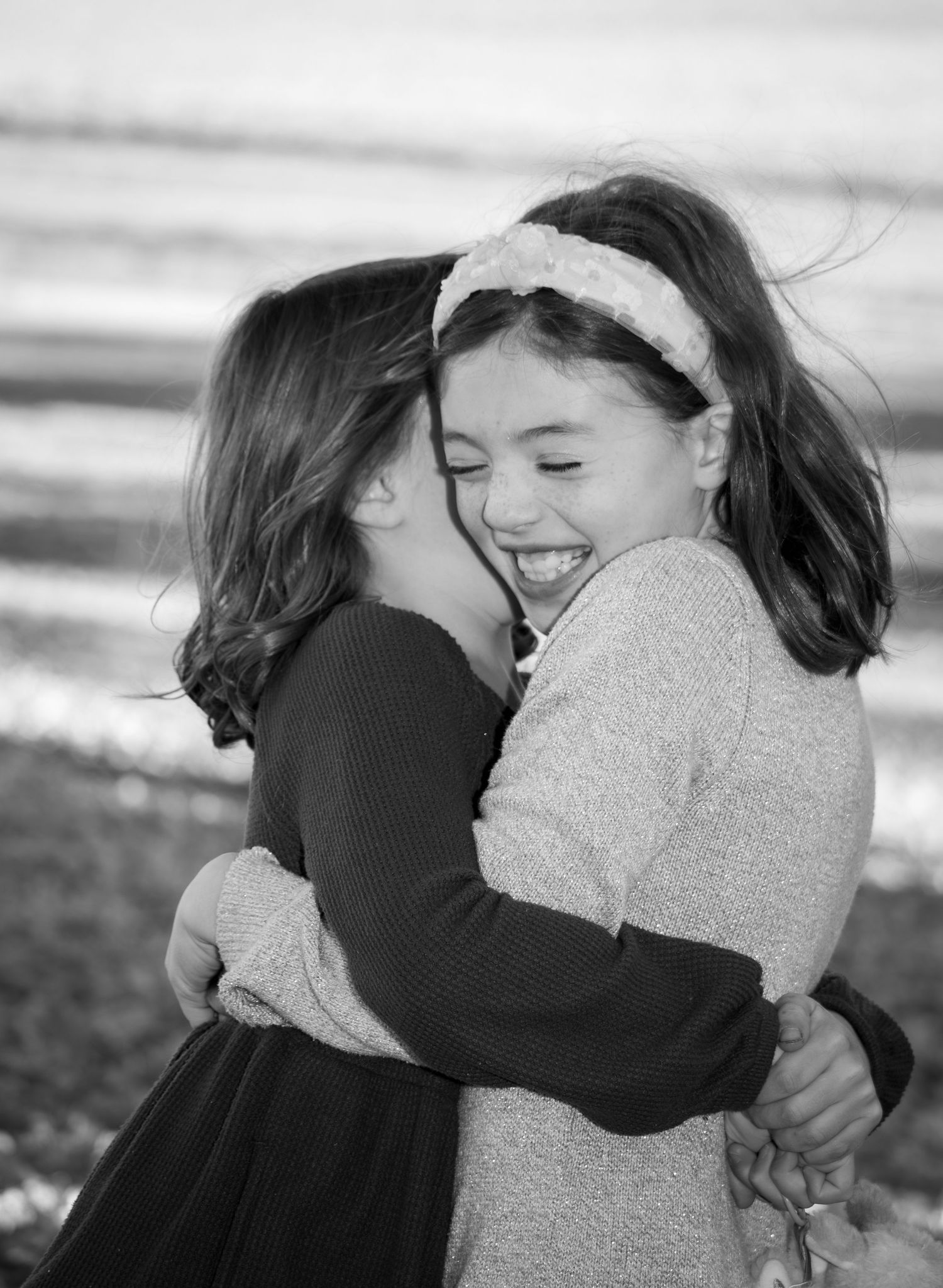 Two young girls share a joyful hug and laugh together in a black and white outdoor photo.
