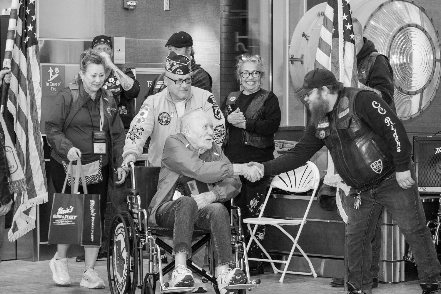 Veteran in wheelchair receiving a handshake at a military honor ceremony with flags displayed.