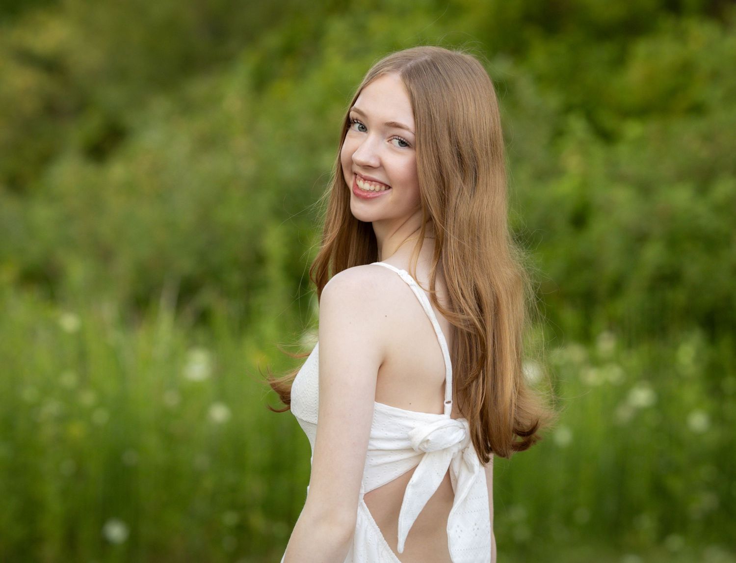 Smiling young woman with long auburn hair wearing a white sundress outdoors in a lush green field.