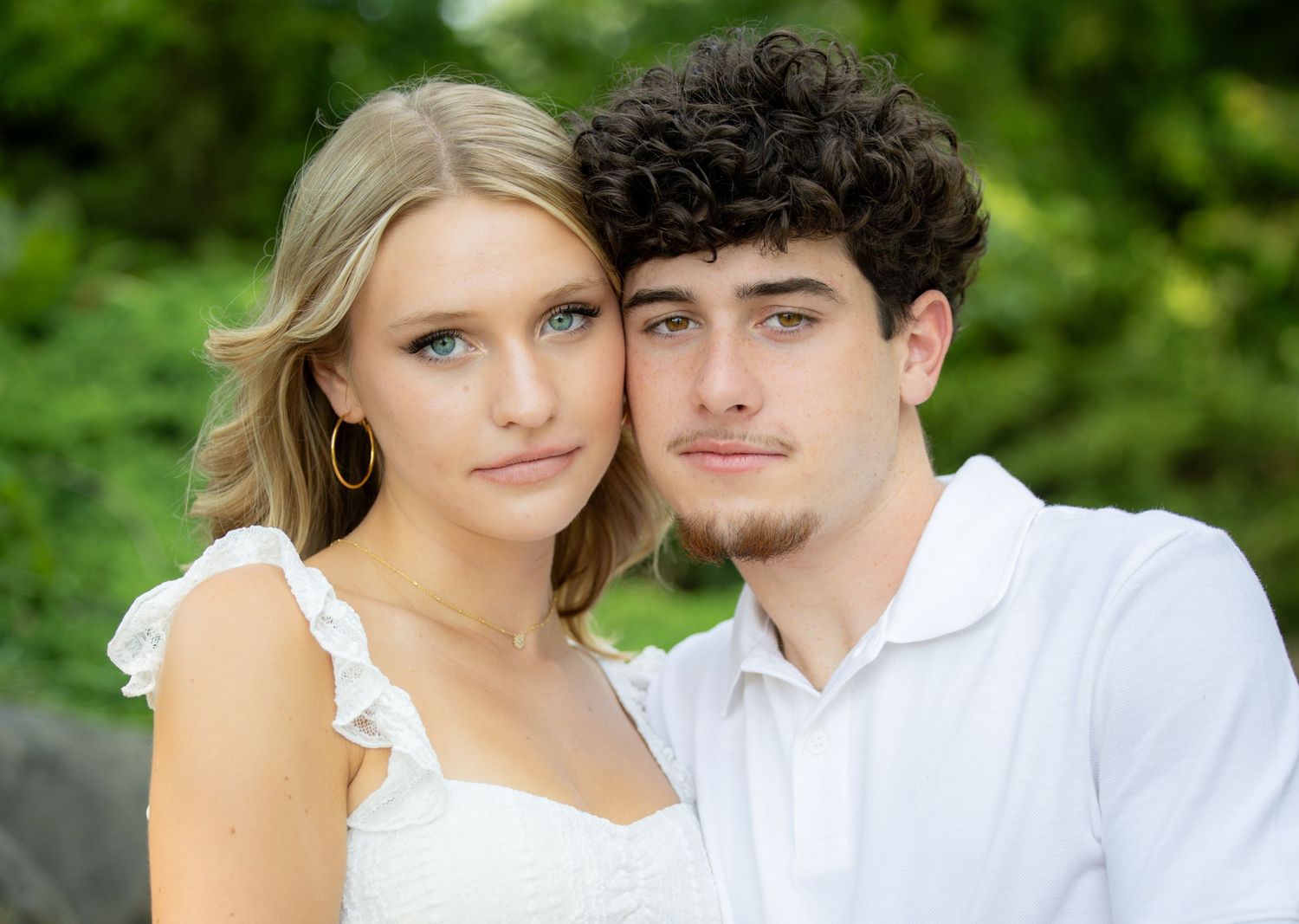 A young couple posing closely together outdoors, woman in white dress with blonde hair, man in white shirt with curly dark hair.