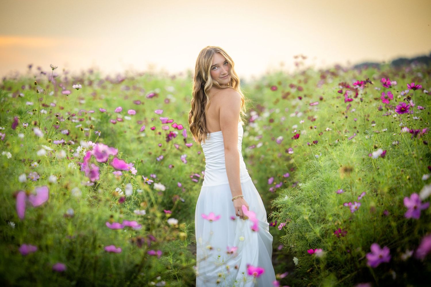 Young woman in white dress standing in a vibrant field of pink cosmos flowers during golden hour.