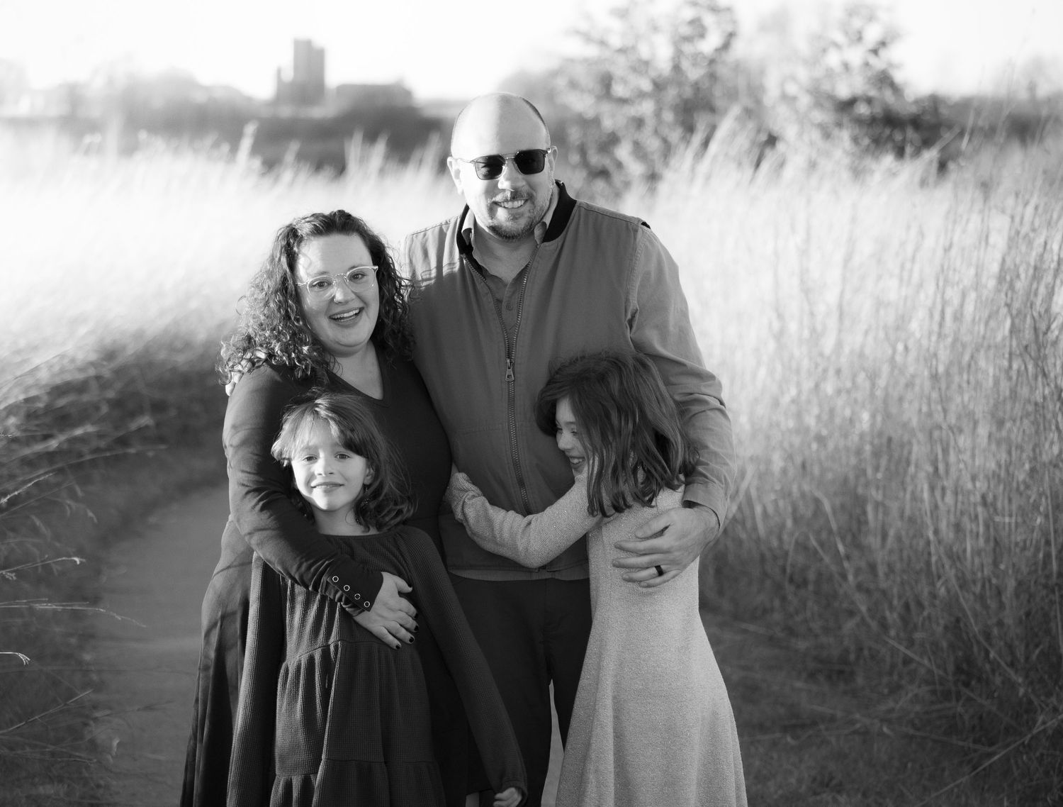 A loving family of four embraces and smiles together on a nature path in black and white.