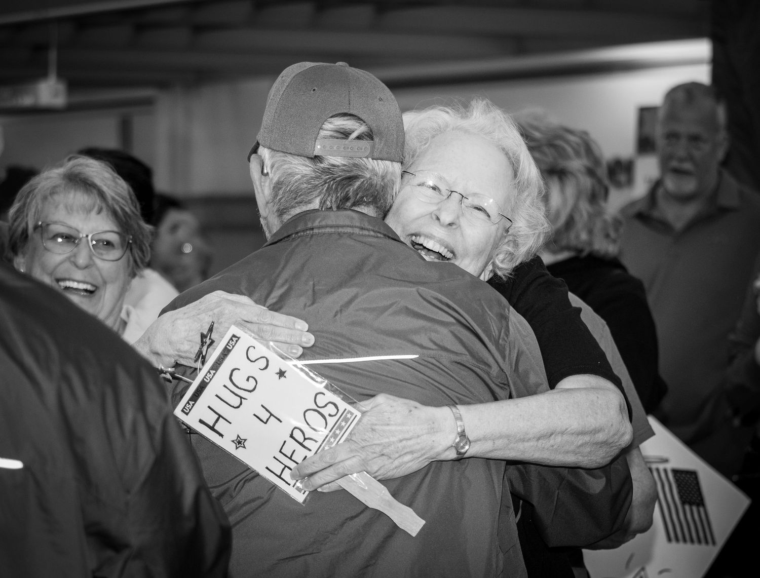 Elderly woman hugging a veteran at a homecoming event holding a Hugs 4 Heroes sign.
