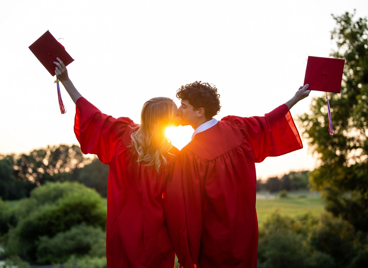 Two graduates in red gowns share a kiss at sunset, holding diplomas aloft in a celebratory outdoor setting.