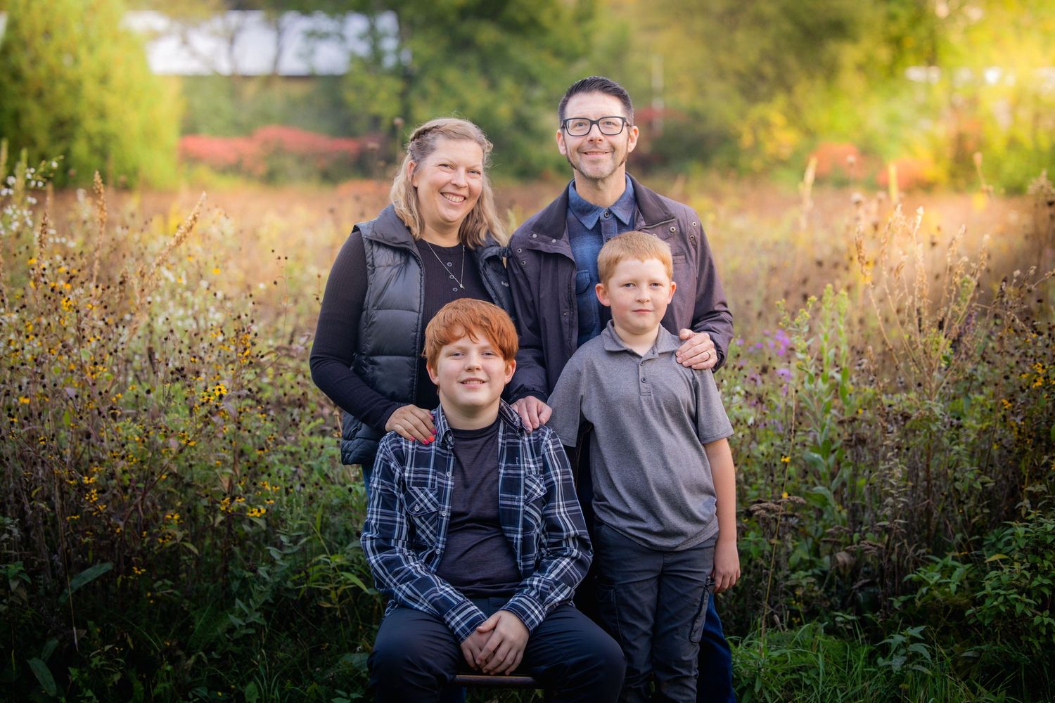 Family of four posing outdoors in a lush green field during golden hour, two adults and two boys smiling.