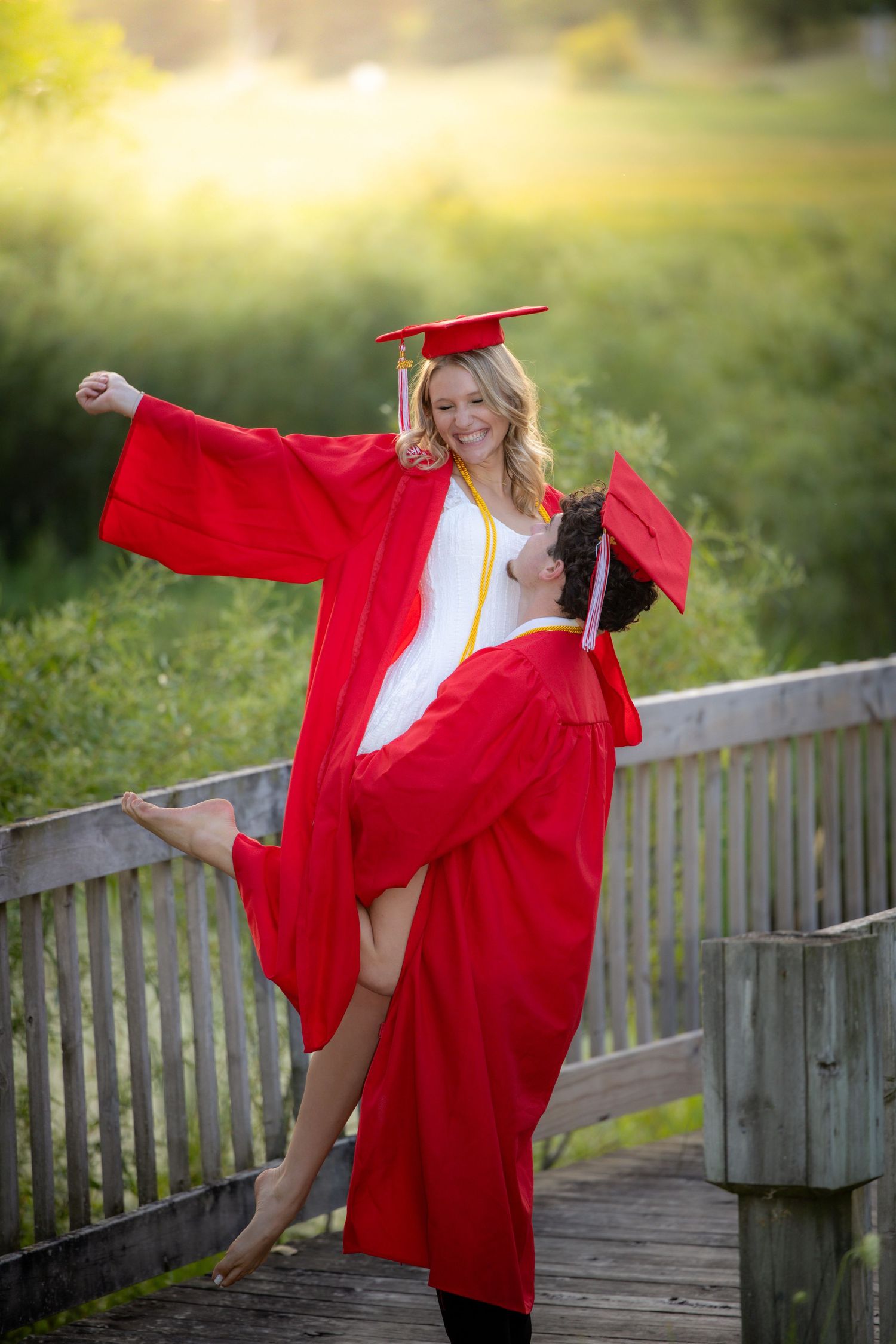 Two graduates in red caps and gowns celebrate joyfully on a wooden bridge surrounded by lush greenery.