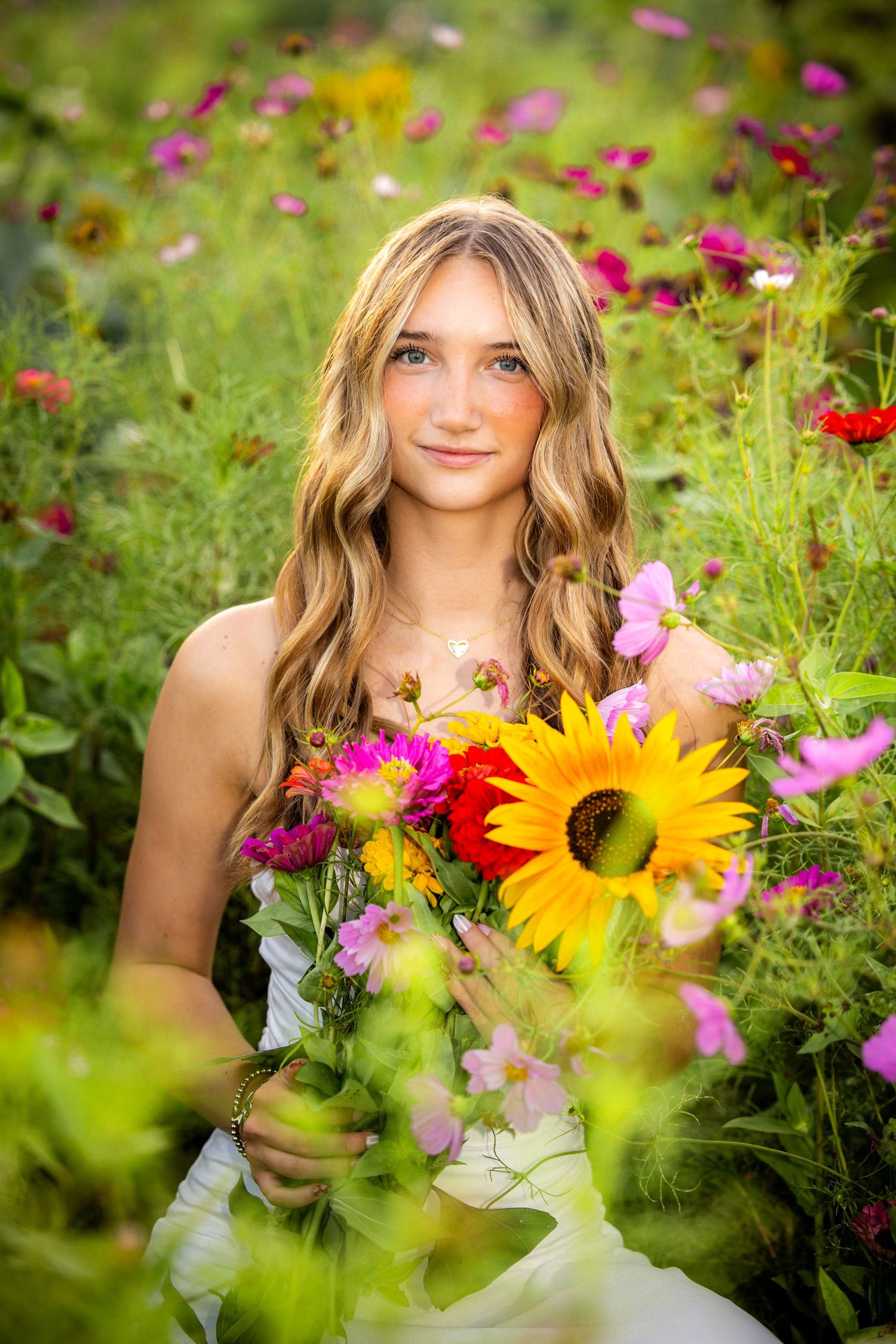 Young blonde woman holding colorful wildflower bouquet with sunflower, sitting in a vibrant garden field.