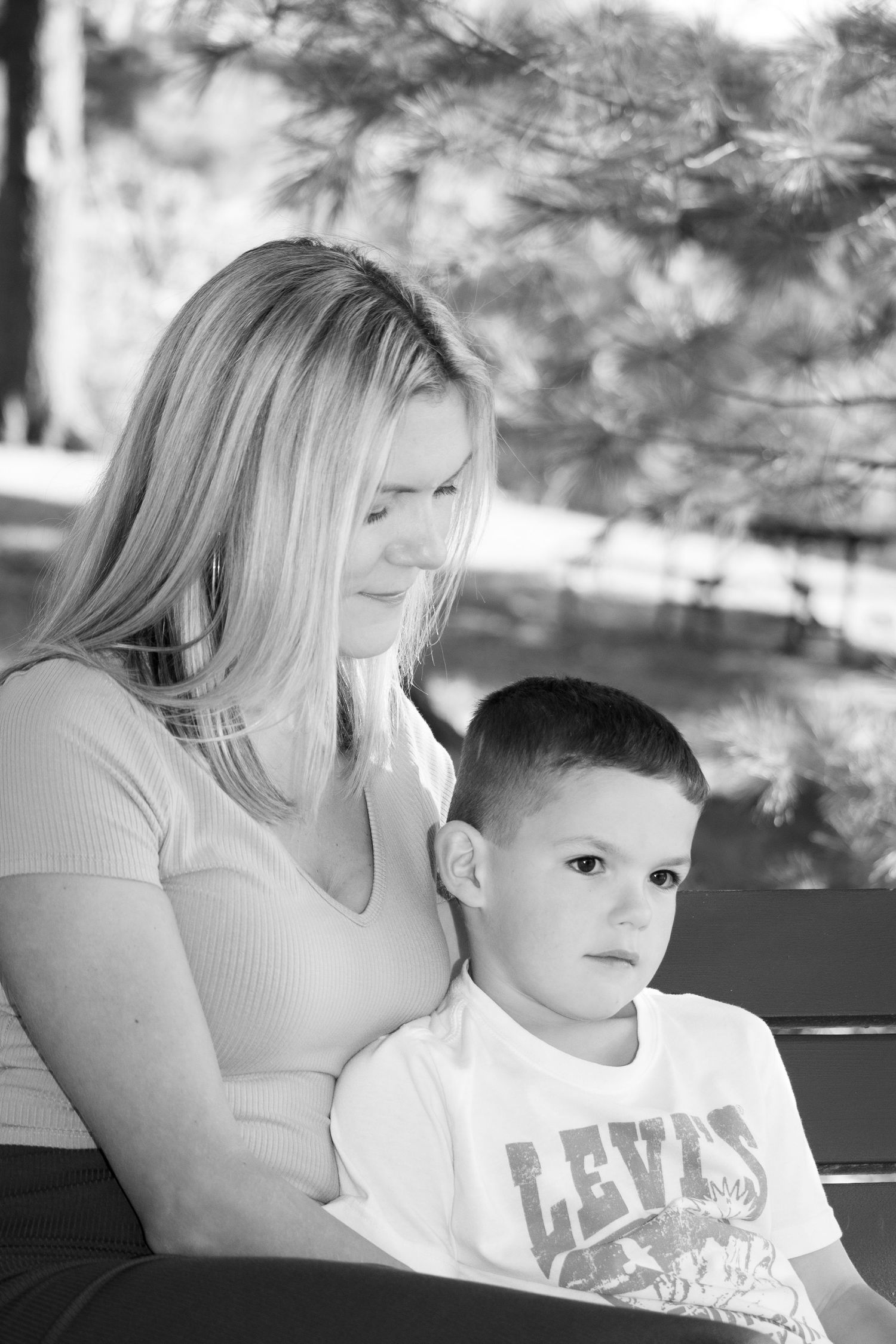 Blonde mother tenderly holds young son on bench in black and white outdoor portrait photo.