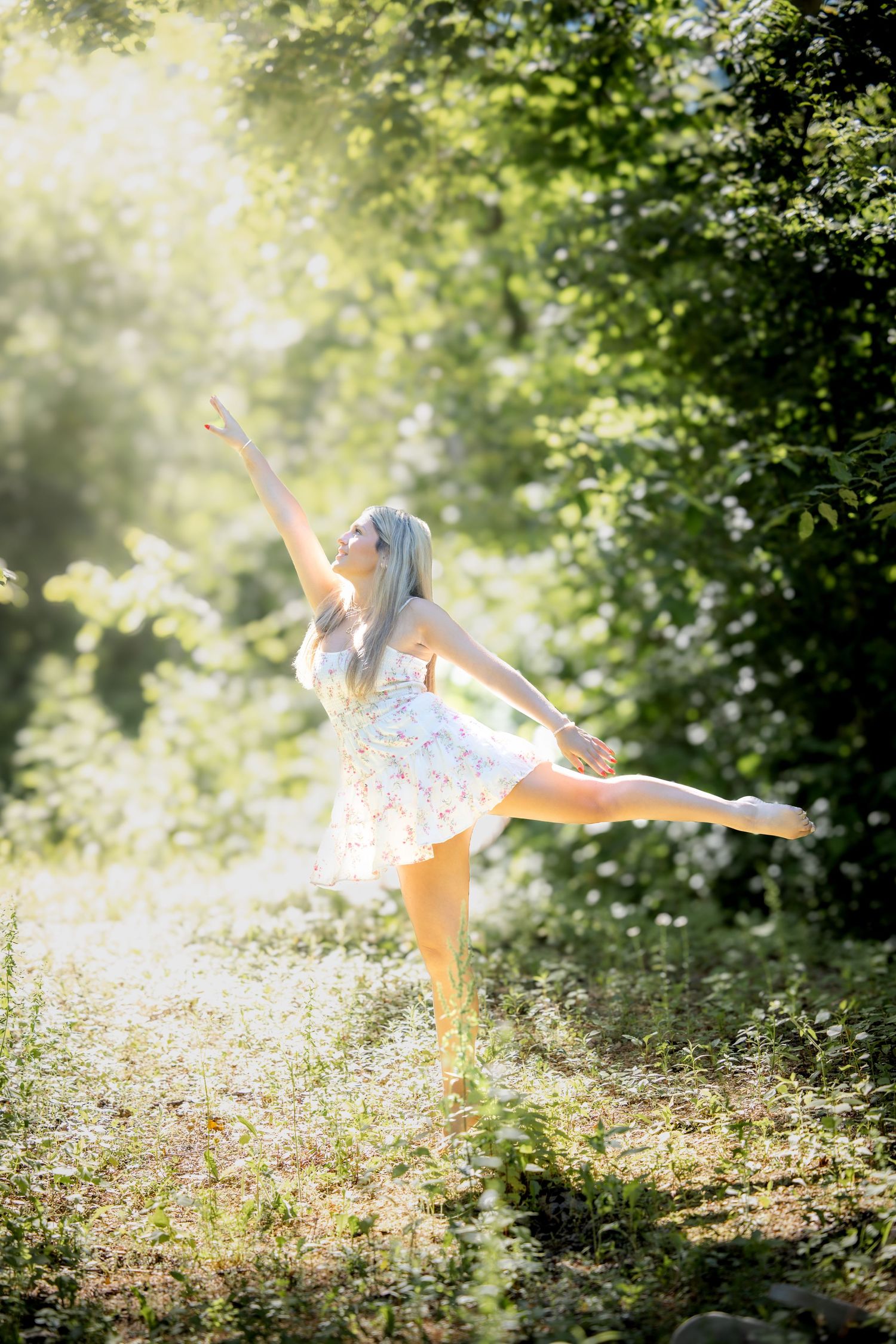 Graceful blonde dancer in floral sundress performing ballet pose in sunlit forest clearing.