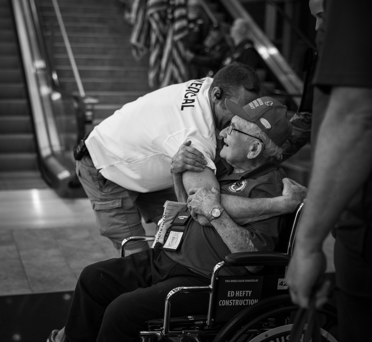 Man leaning down to embrace elderly veteran in wheelchair at indoor public space.