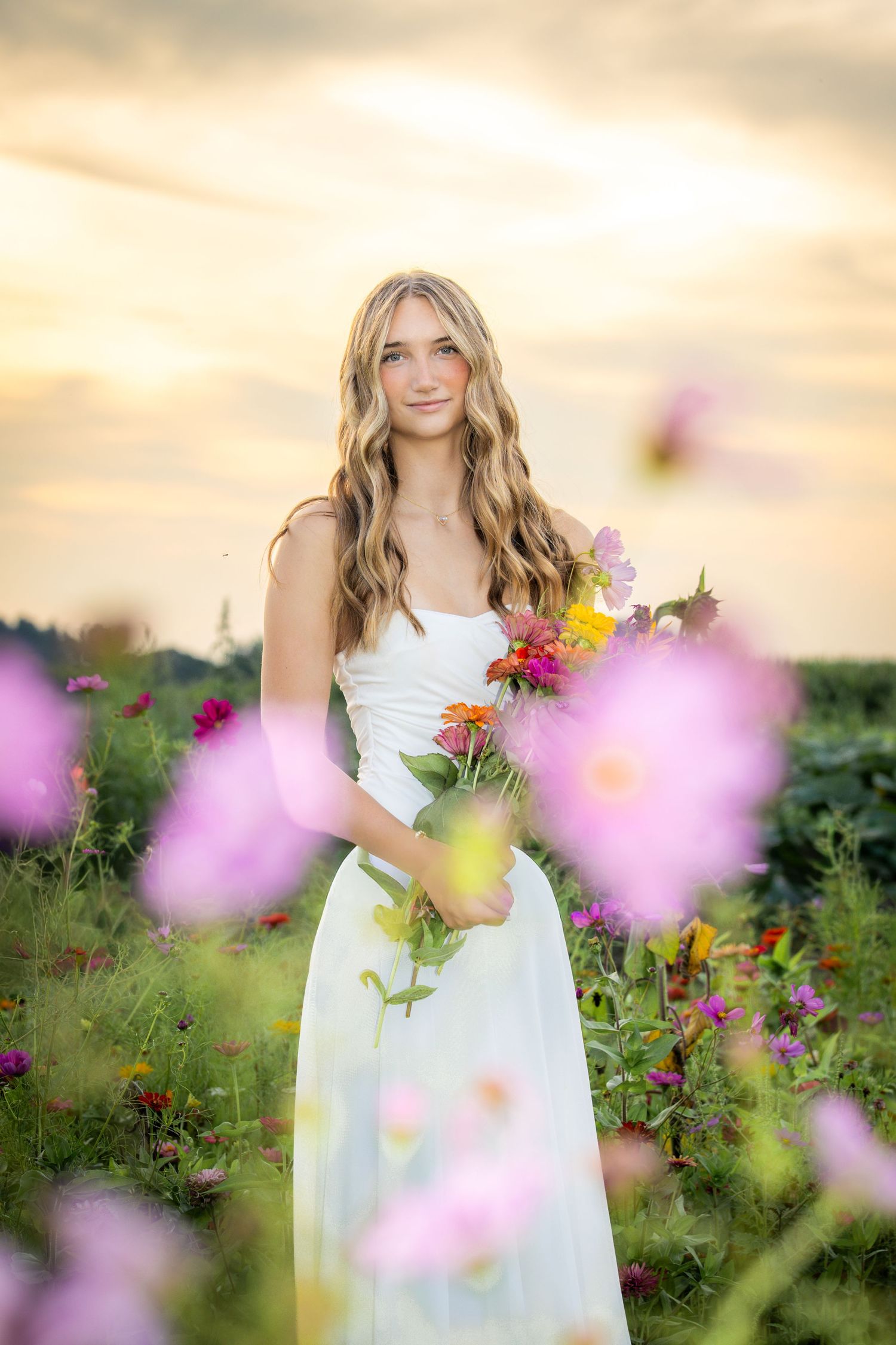 Young woman in white dress holding colorful flowers in a blooming field at golden sunset.