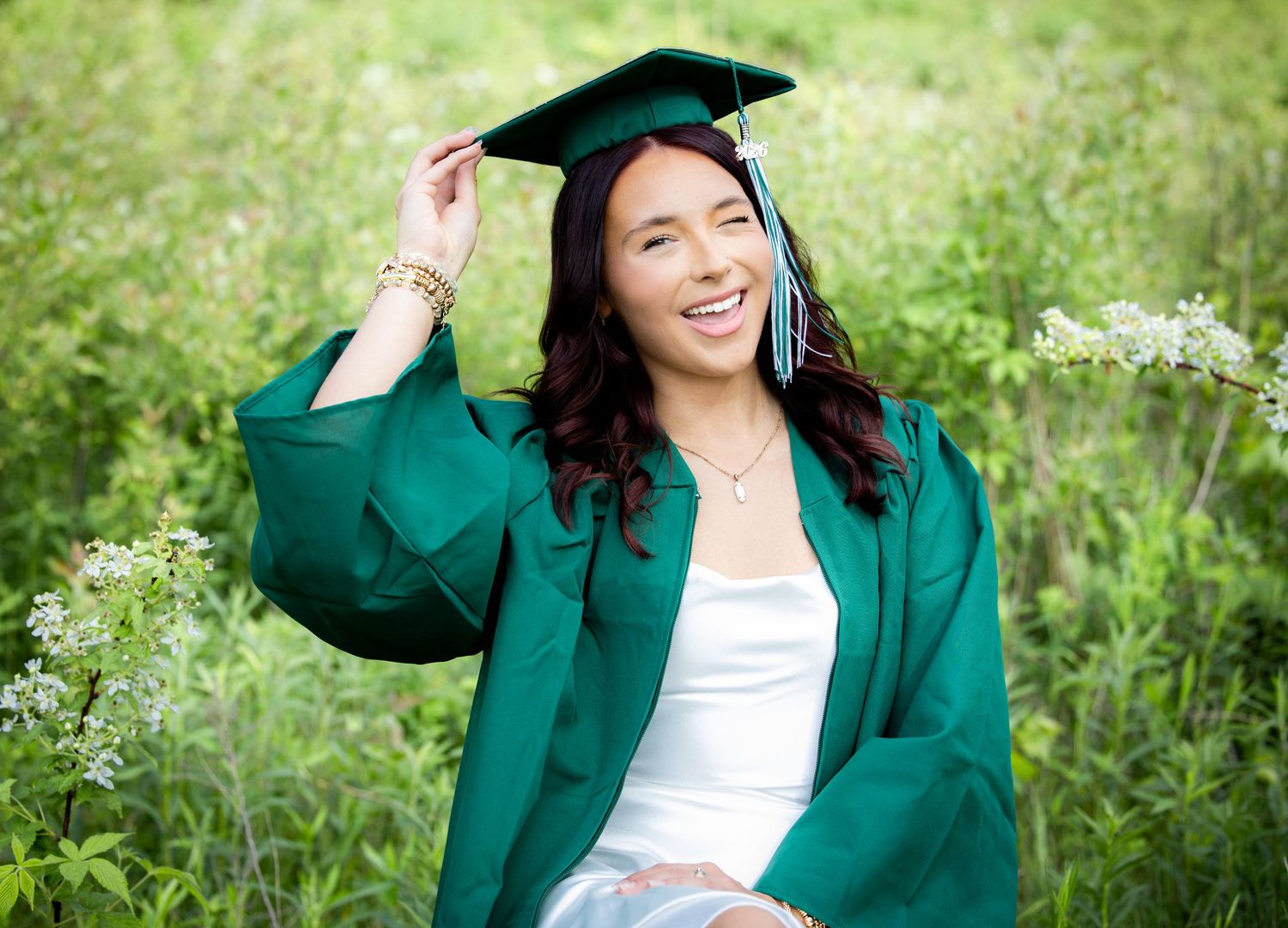 Smiling graduate in green cap and gown poses outdoors in a wildflower field on a bright sunny day.