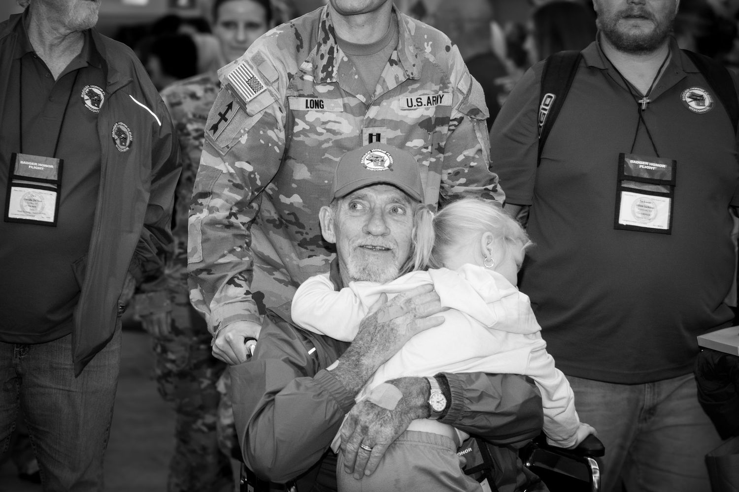 Black and white photo of elderly veteran holding baby surrounded by people at indoor event.