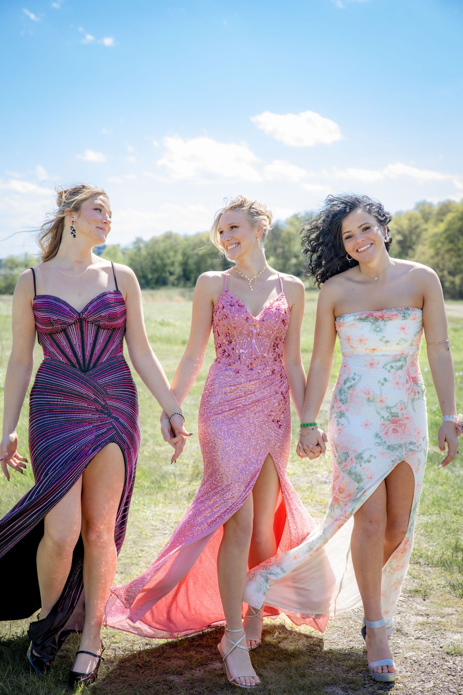 Three young women in elegant prom dresses walk hand-in-hand through a sunny field, smiling joyfully.