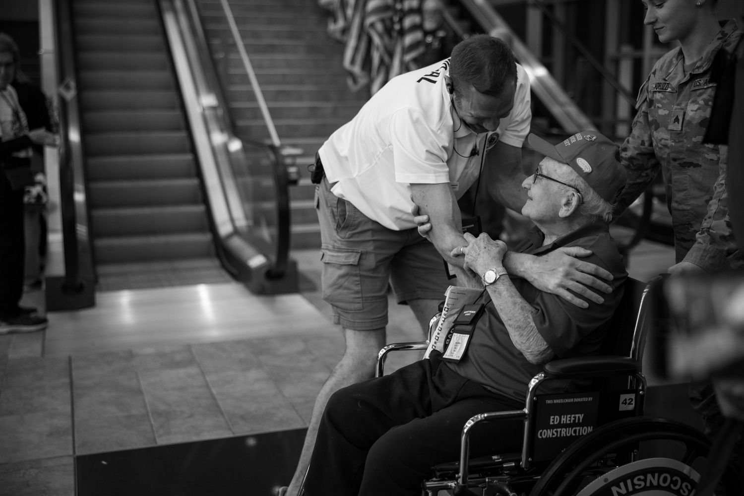 Man leaning down to embrace a veteran in a wheelchair inside a building with an escalator.