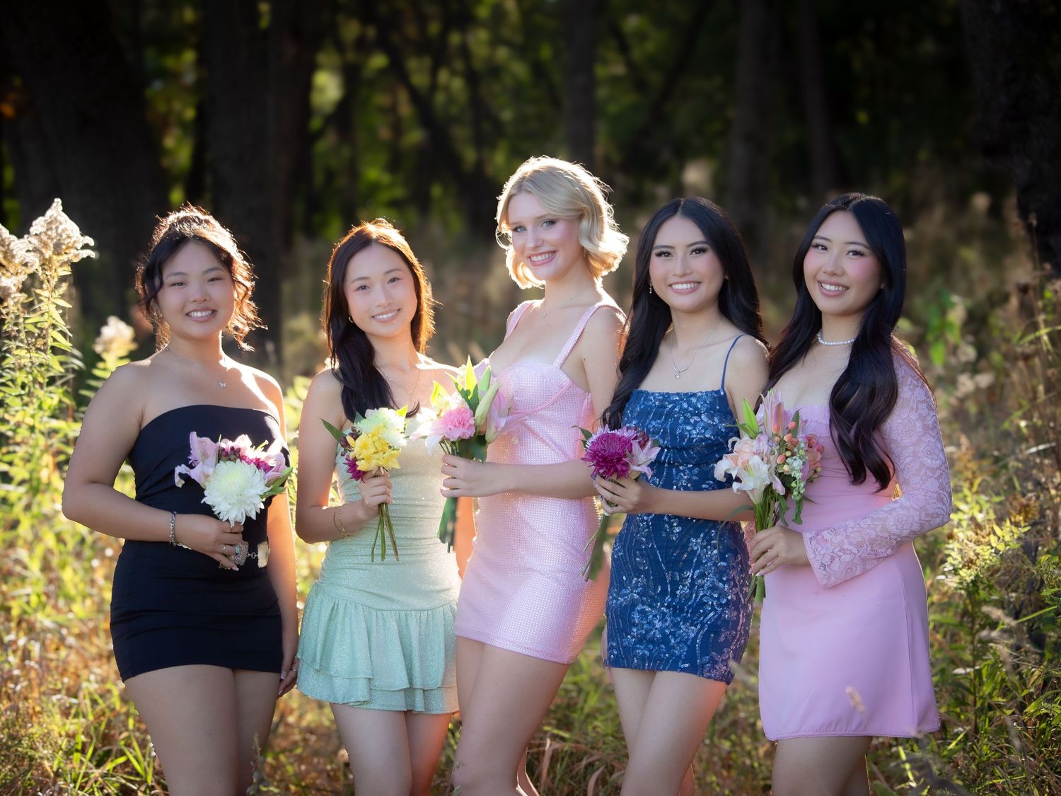 Five young women in colorful formal dresses holding flowers posing together in a forest setting.